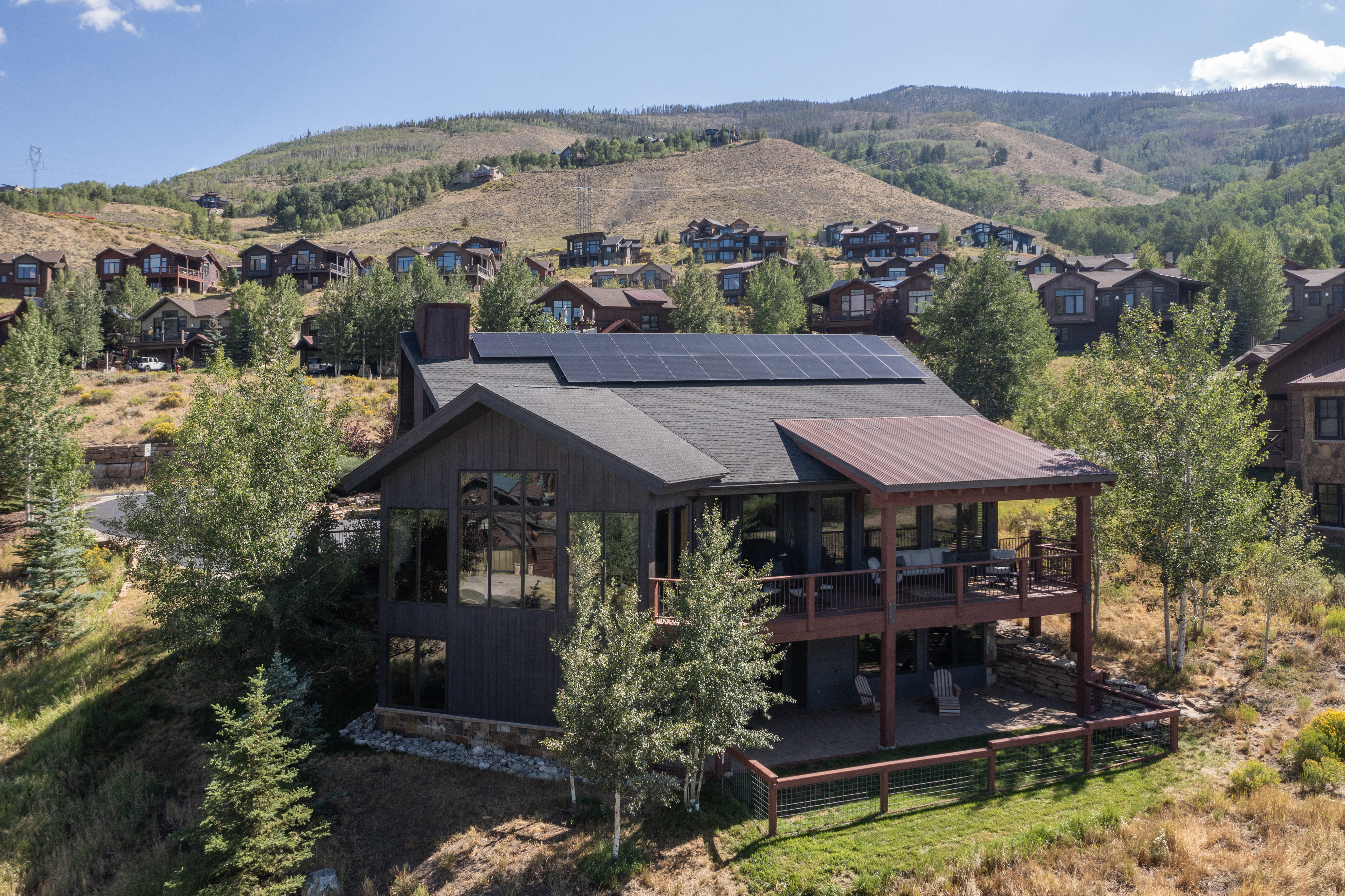 This elevated perspective captures a modern mountain home nestled into a hillside, featuring dark wood siding and a prominent roofline equipped with solar panels. The residence includes a spacious elevated deck and a lower-level stone patio, both overlooking a natural, grassy landscape with scattered aspen trees. The surrounding terrain is characterized by rolling hills and a community of similar mountain-style homes under a clear blue sky.