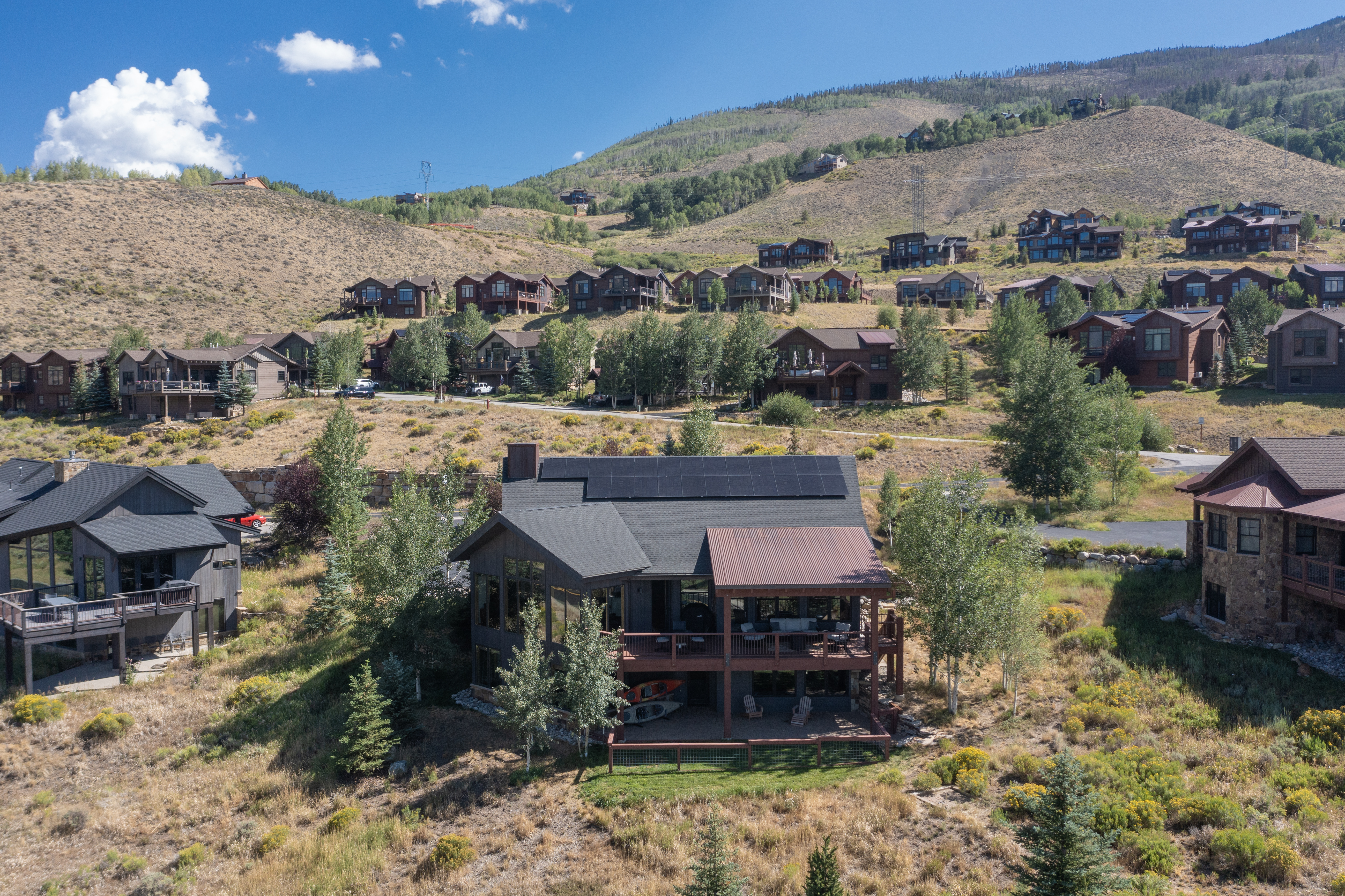 This elevated aerial perspective captures a mountain-side residential community featuring several multi-story homes with dark wood siding and shingled roofs. The central focus is a prominent residence with a large solar panel array on its roof and a spacious multi-level deck overlooking the rugged, dry landscape. The surrounding terrain is characterized by rolling hills, scattered evergreen trees, and a clear blue sky, emphasizing the secluded and scenic nature of the neighborhood.