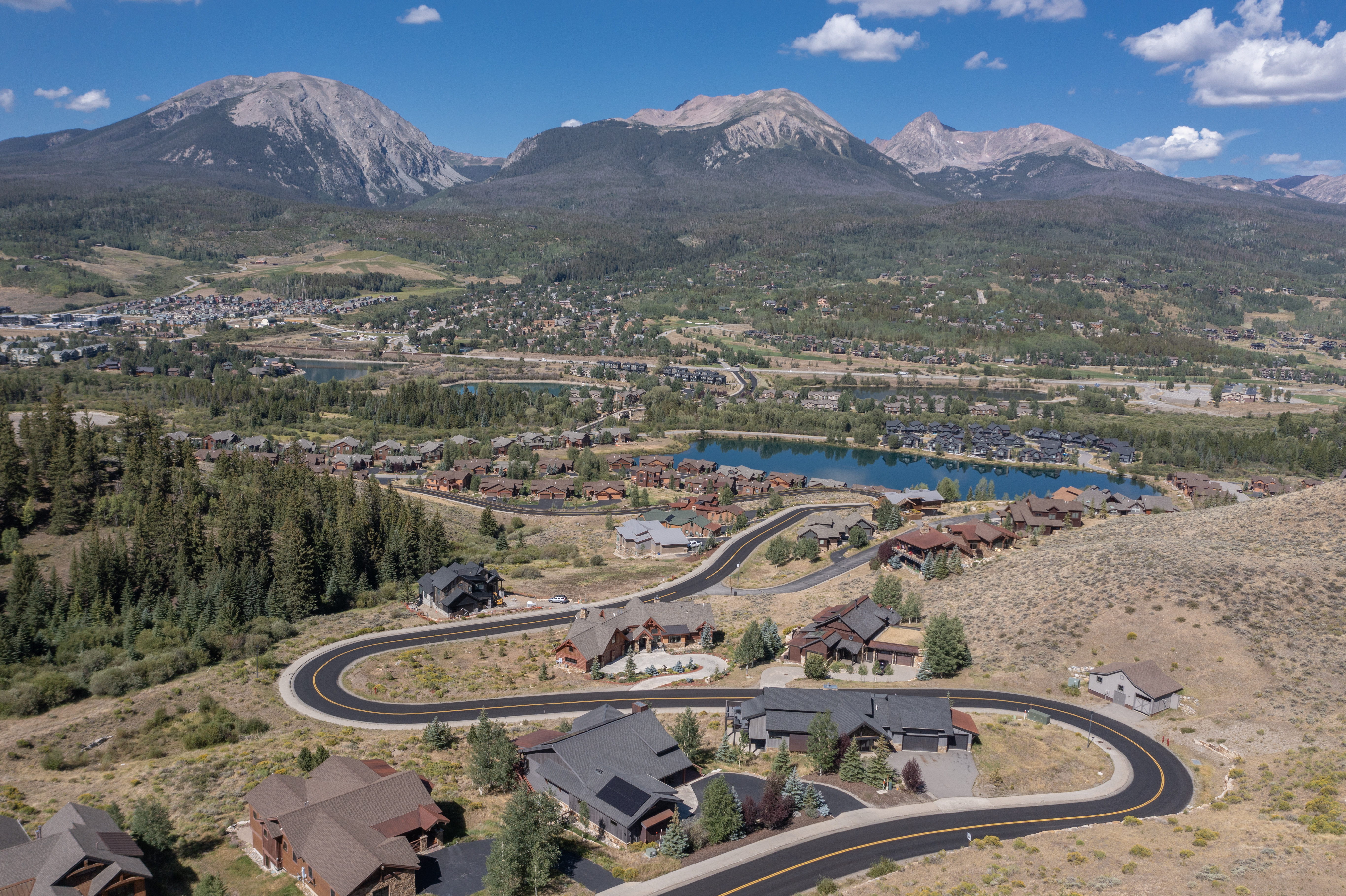 This high-angle aerial view captures a sprawling mountain community nestled in a valley beneath dramatic, rugged peaks. The scene features winding paved roads connecting large, custom-built homes with dark-toned roofs, surrounded by a mix of evergreen trees and dry, golden-hued terrain. A serene blue lake sits centrally in the valley, providing a striking contrast to the surrounding landscape and emphasizing the scenic, secluded nature of the neighborhood.