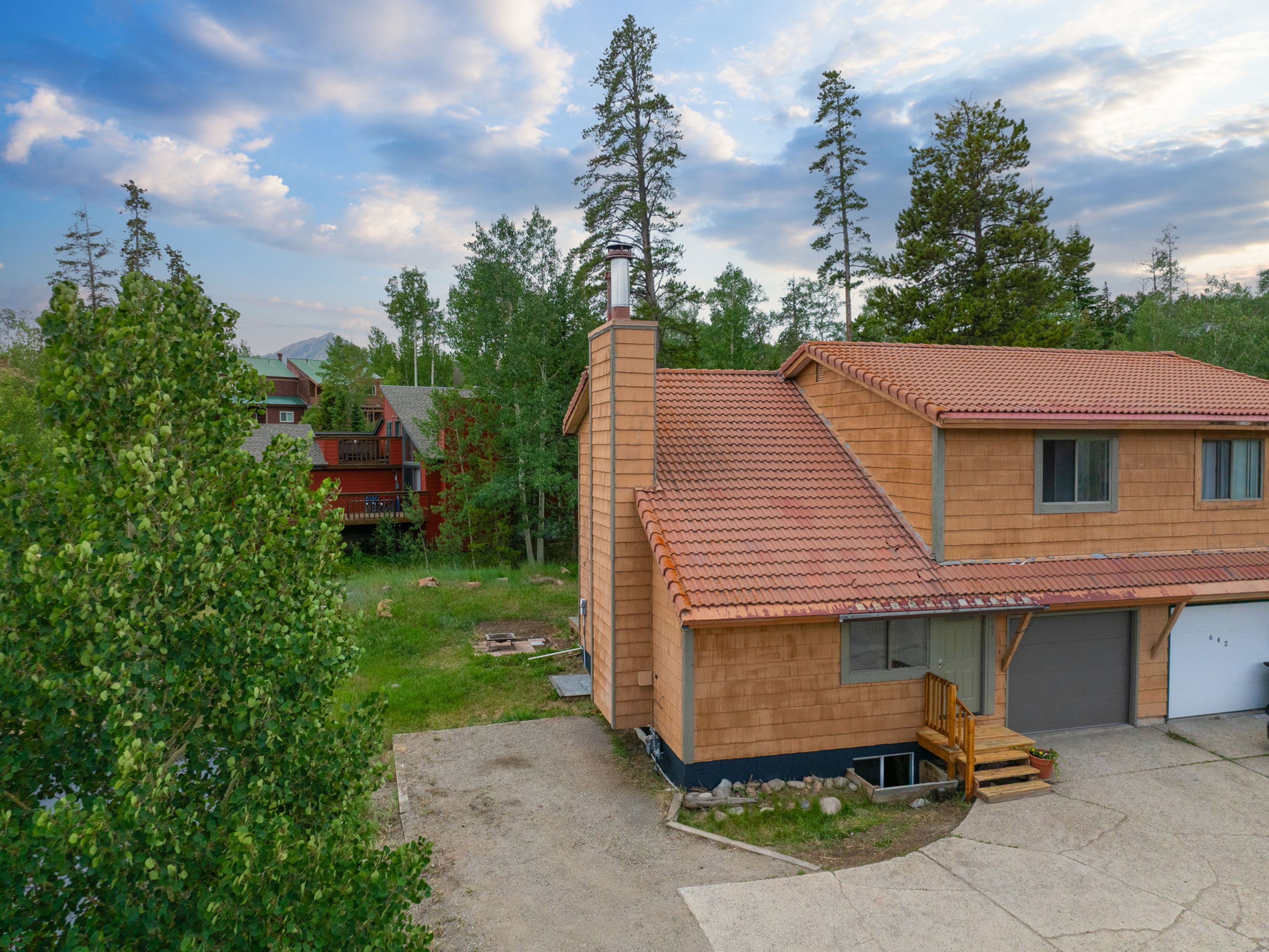 This elevated perspective captures the front exterior of a multi-level home featuring warm-toned wood siding and a prominent chimney structure. The property includes a concrete driveway leading to a garage, a small wooden staircase leading to the entrance, and a surrounding landscape of mature trees and greenery. The scene is set under a soft, cloudy sky, highlighting the home's rustic mountain aesthetic.