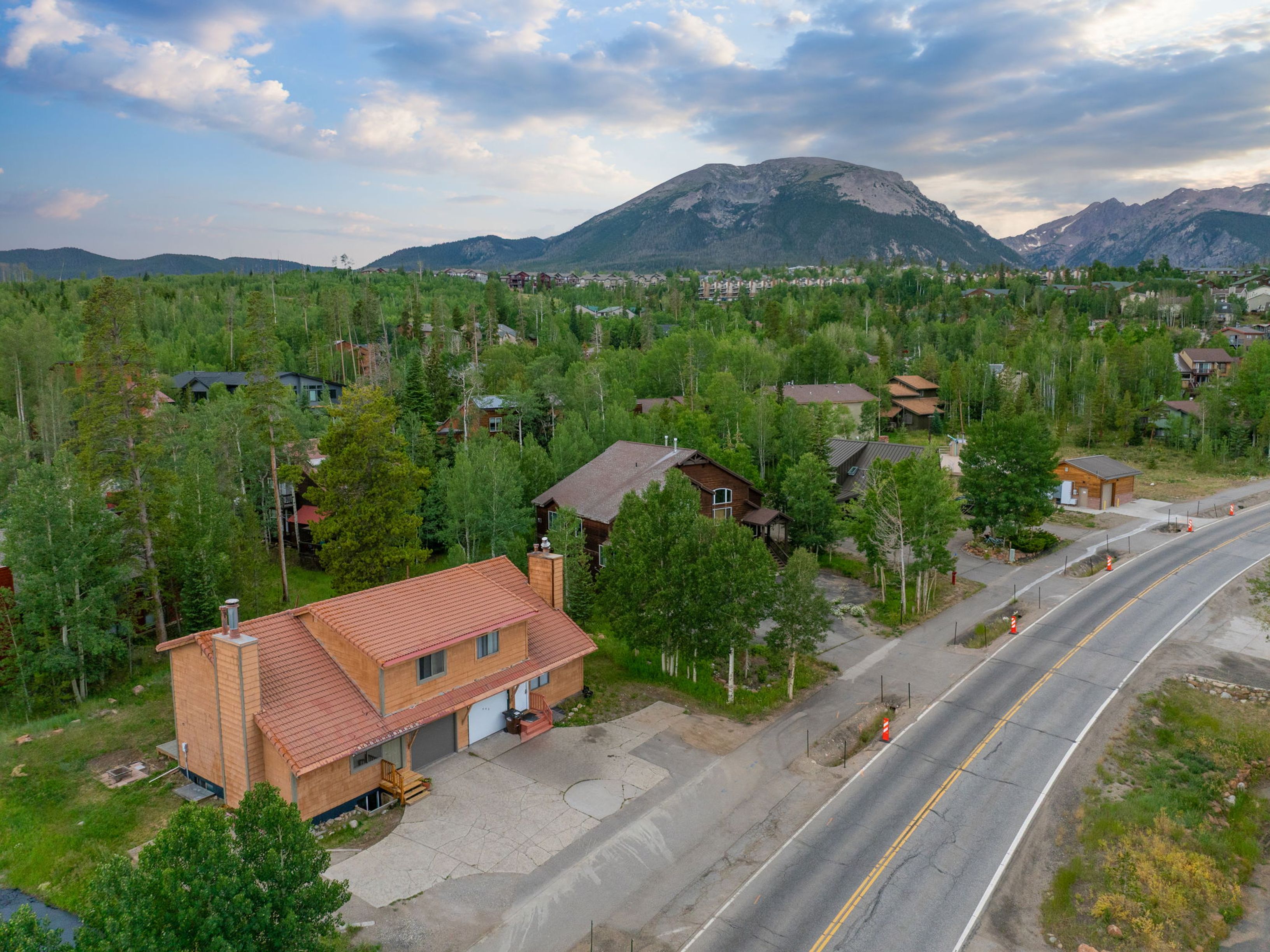 This high-angle aerial view captures a multi-level home with a distinctive reddish-brown tiled roof, situated in a lush, forested mountain setting. The property features a paved driveway, a two-car garage, and a prominent chimney structure, all set against a backdrop of dense pine trees and a majestic mountain peak under a cloudy sky. The perspective highlights the home's integration into the natural landscape and its proximity to a winding road.