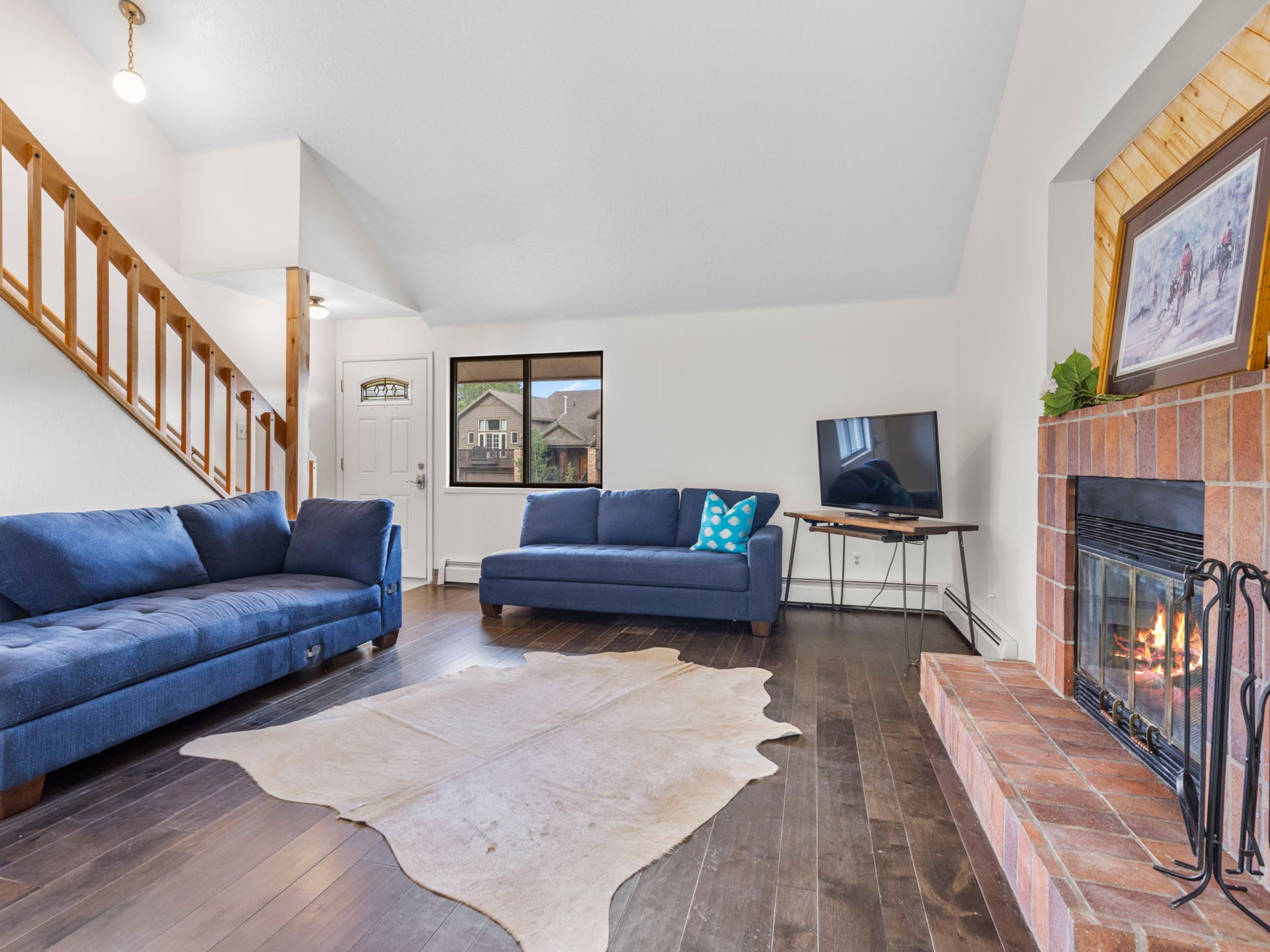 This bright living room features dark wood flooring, a vaulted ceiling, and a prominent brick fireplace with a hearth. Two blue sofas are arranged in the space, complemented by a cowhide rug and a television set on a minimalist stand. A staircase with a wooden railing leads upward, and a window provides natural light, creating an open and inviting atmosphere.