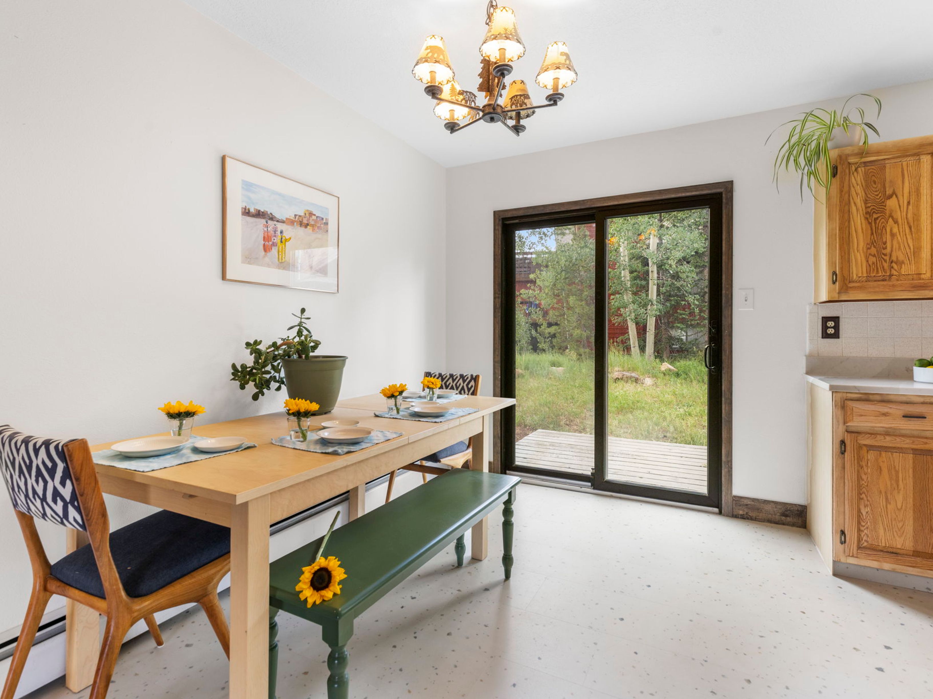 This bright dining area features a light wood table paired with a mix of patterned chairs and a green painted bench. A large sliding glass door provides a clear view of the outdoor deck and wooded yard, while a decorative chandelier hangs above the table. The space is finished with light-colored flooring and adjacent wooden kitchen cabinetry, creating a casual and inviting atmosphere.