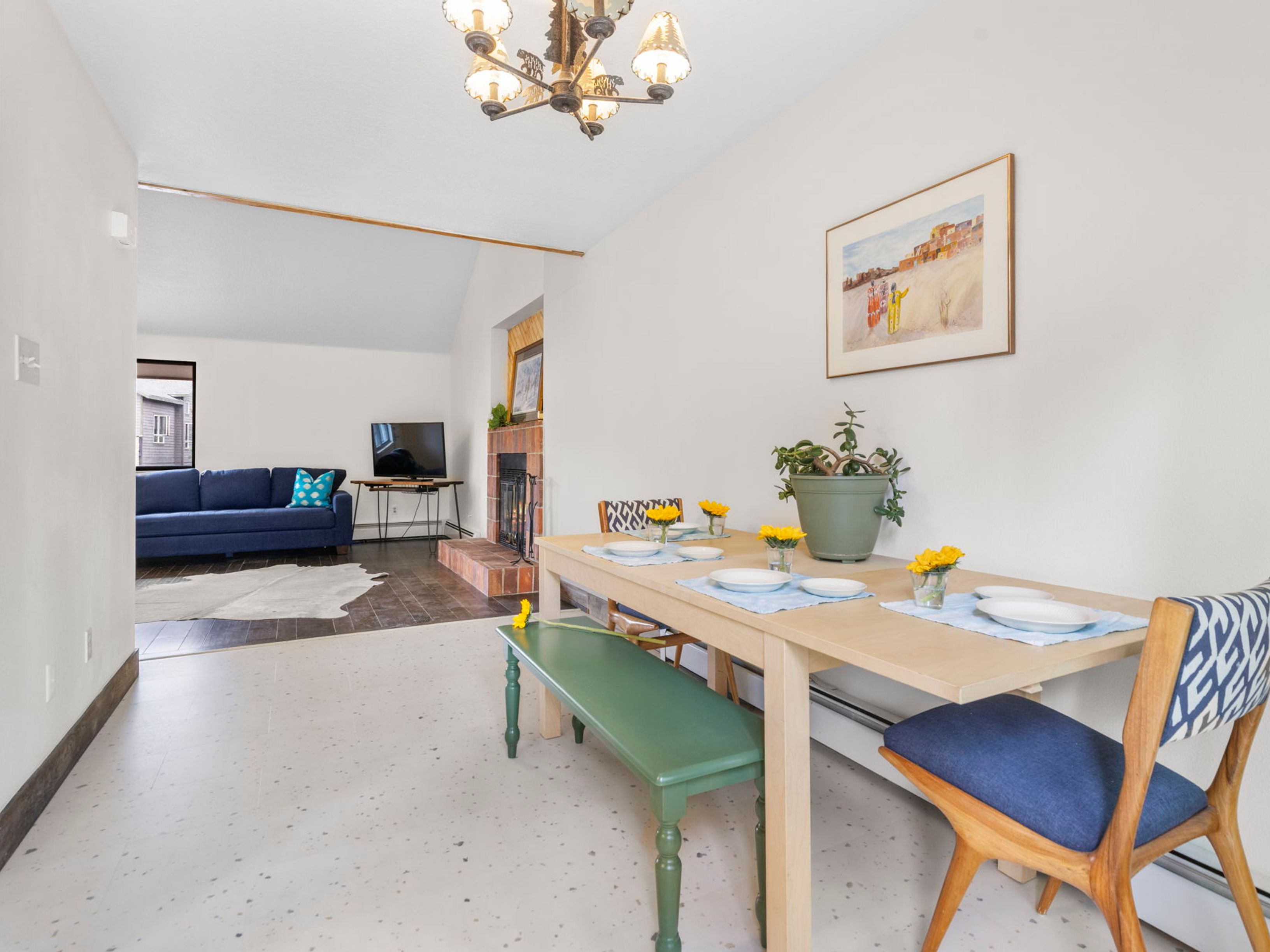 This open-concept living and dining area features a light wood dining table set for four, accompanied by a green bench and patterned chairs. Beyond the dining space, a step-down living room showcases a blue sofa, a fireplace with a brick surround, and a television on a metal stand. The home is characterized by a neutral color palette, speckled flooring, and a decorative chandelier, creating a bright and inviting atmosphere.
