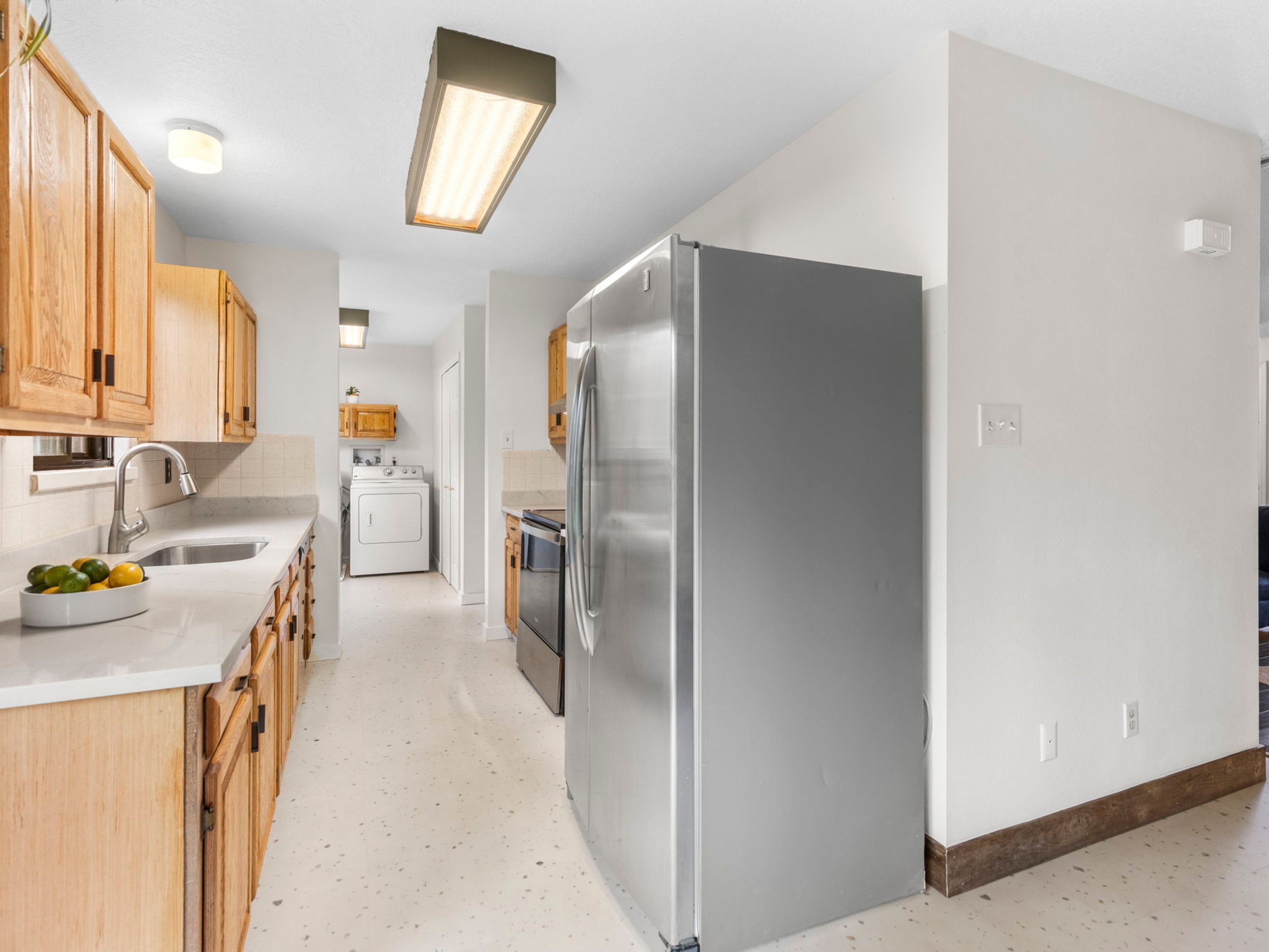 This galley-style kitchen features light wood cabinetry, a stainless steel refrigerator, and a neutral countertop with an integrated sink. The space includes a light-colored floor with subtle speckling and leads directly into a laundry area equipped with a white washer and dryer. The perspective is a straight-on shot looking down the length of the kitchen, highlighting the functional layout and transition between the cooking and utility zones.