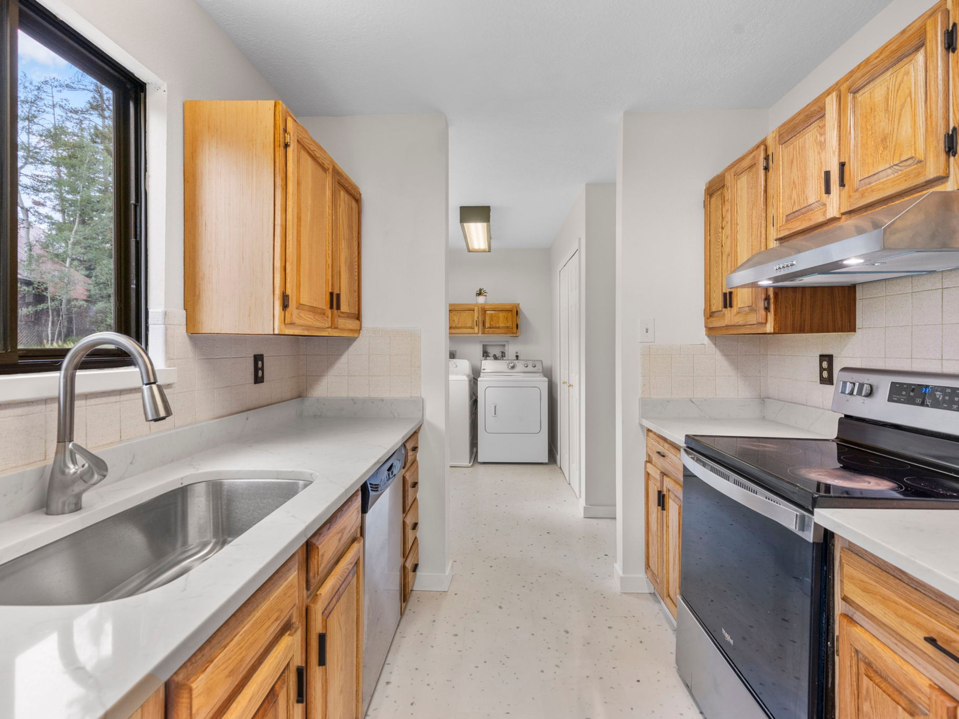 This galley-style kitchen features light wood cabinetry paired with light-toned stone countertops and a stainless steel sink. The space includes a black electric range with a matching vent hood and a dishwasher, leading directly into a laundry area with a washer and dryer. A window above the sink provides natural light, while the neutral color palette creates a clean and functional atmosphere.
