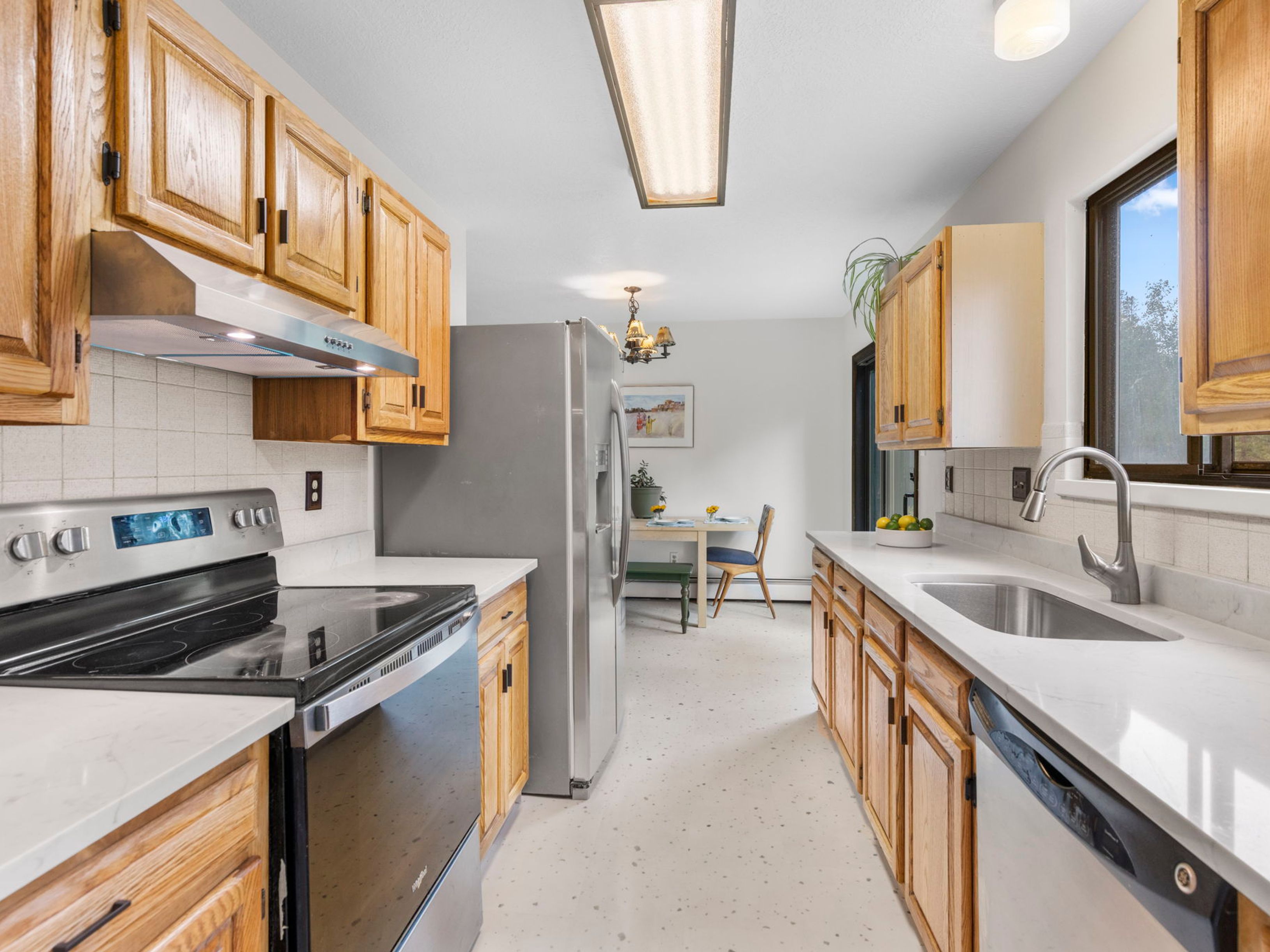 This galley-style kitchen features light wood cabinetry, white countertops, and stainless steel appliances, including a range and a refrigerator. The space is illuminated by a long, rectangular overhead light fixture and includes a sink positioned beneath a window. A view into the adjacent dining area reveals a table with seating, creating an open and functional flow between the cooking and eating spaces.