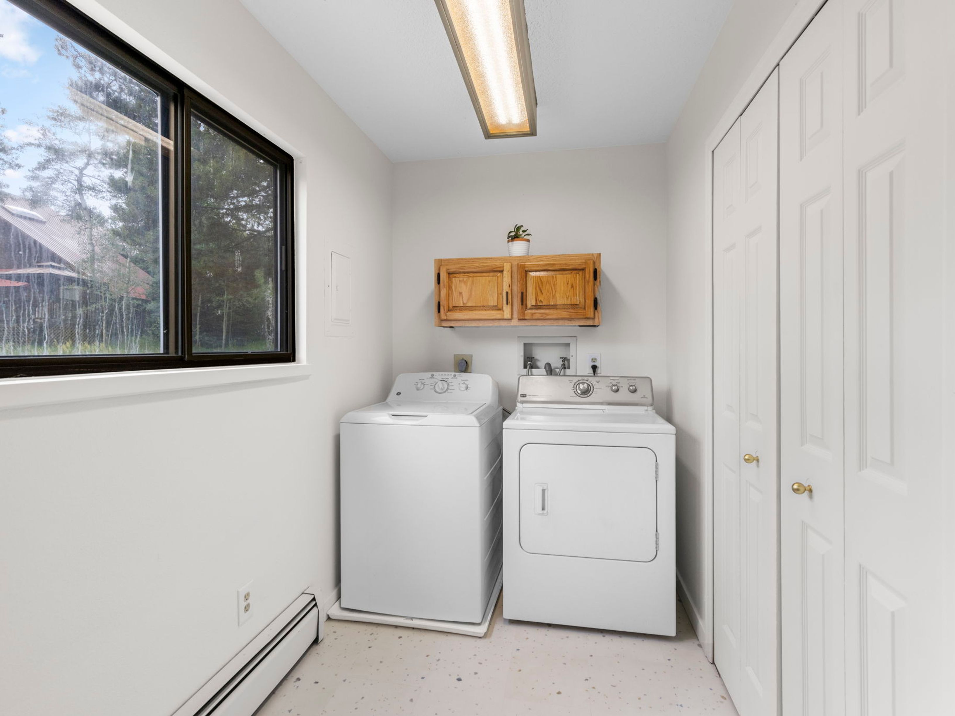 This functional laundry room features a side-by-side washer and dryer set positioned against a neutral-toned wall. Above the appliances, a light wood wall-mounted cabinet provides storage, while a large window to the left allows natural light to brighten the space. The room is finished with simple flooring and a standard ceiling-mounted light fixture, offering a clean and practical utility area.