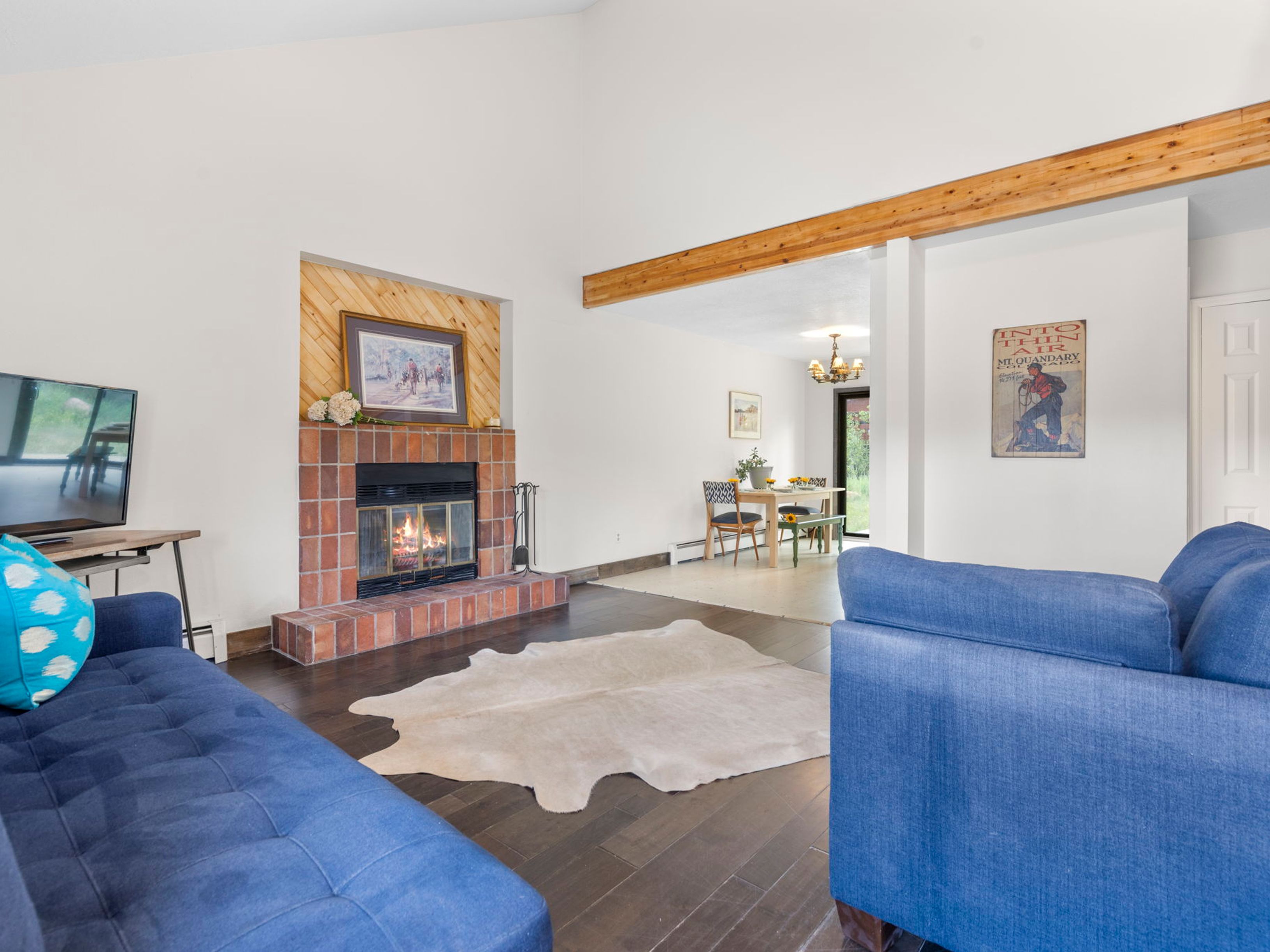 This open-concept living room features dark wood flooring and a prominent brick fireplace with a decorative wood-paneled accent wall above it. Two vibrant blue sofas frame a central cowhide rug, creating a cozy seating area that flows directly into an adjacent dining space. A large wooden beam spans the ceiling, adding architectural character to the bright, airy room.