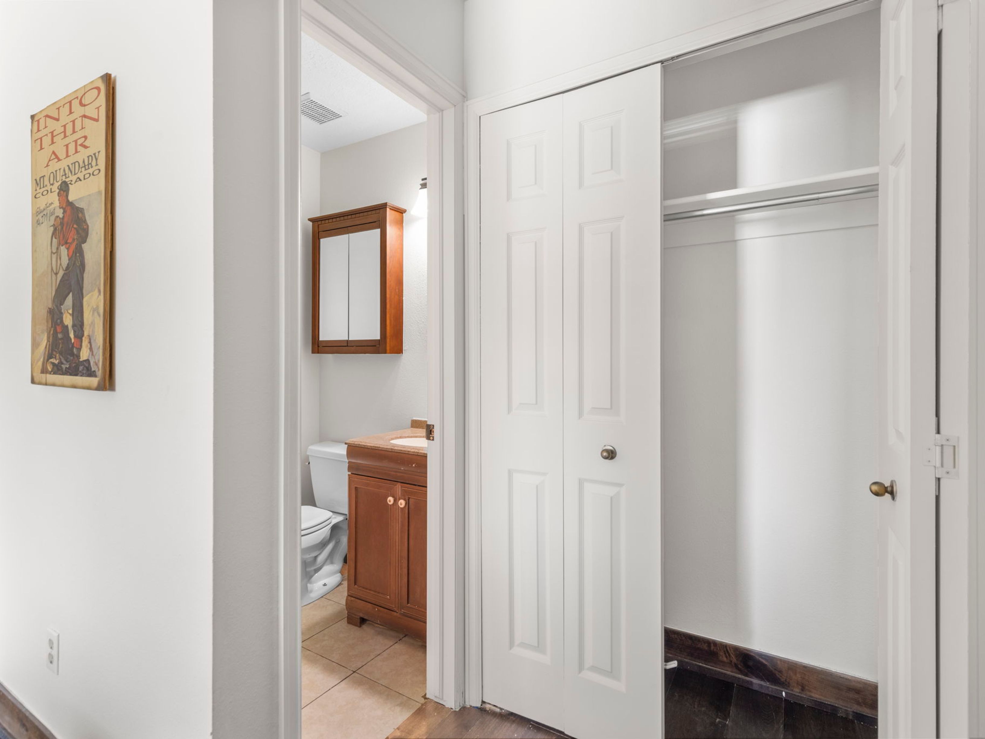 This view captures a transition space featuring a hallway that leads into a bathroom and an adjacent closet with white bi-fold doors. To the left, a framed vintage-style poster hangs on a neutral wall, while the flooring transitions from dark wood in the foreground to light tile inside the bathroom. The bathroom is equipped with a wooden vanity, a medicine cabinet, and a standard toilet, creating a functional and compact layout.