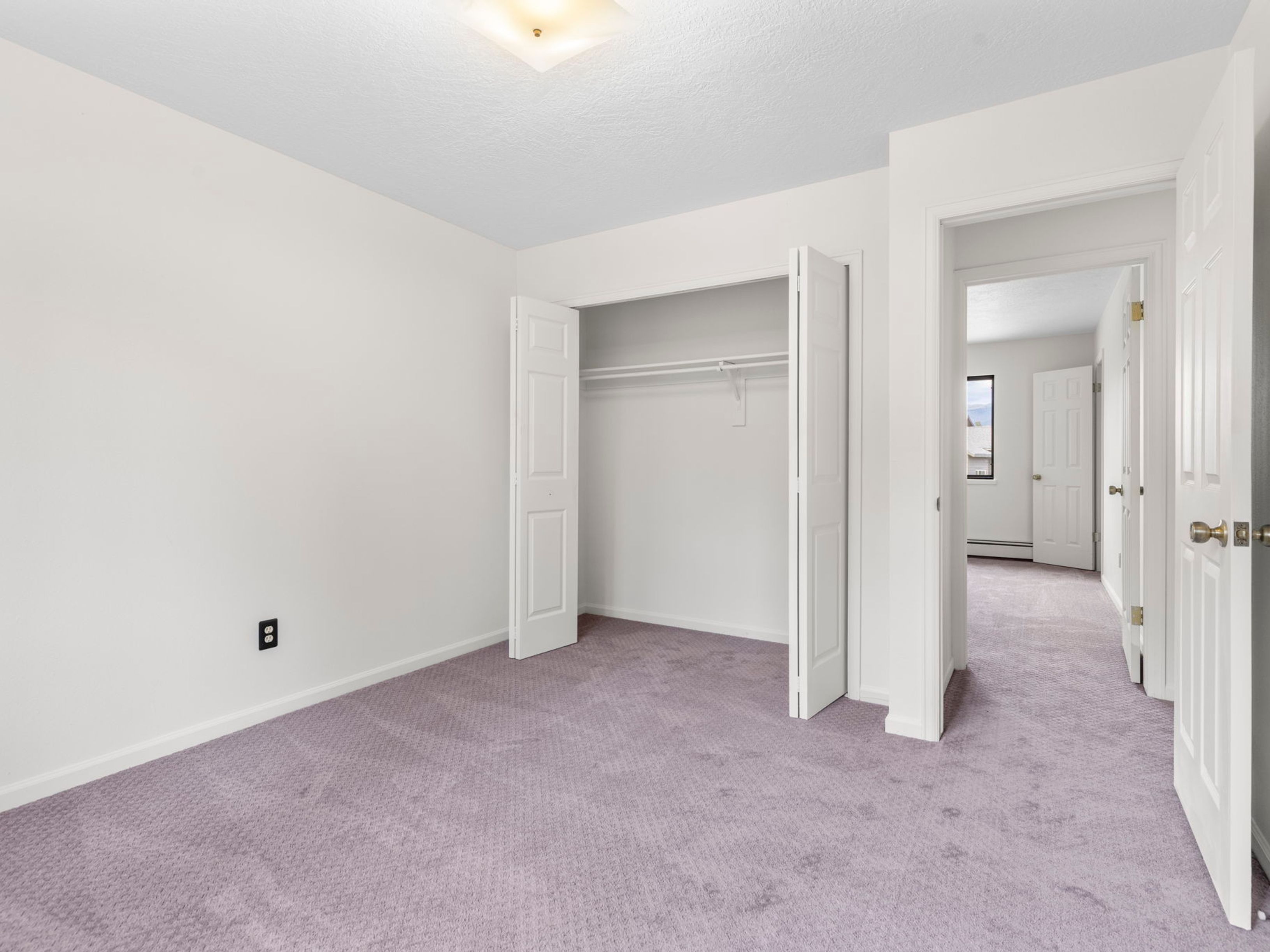 This bright, neutral-toned bedroom features light-colored walls and wall-to-wall purple carpeting. The room includes a built-in closet with white bi-fold doors and a doorway leading into an adjacent hallway, providing a sense of flow between spaces. The perspective is from the corner of the room, capturing the clean, minimalist aesthetic and the simple overhead lighting fixture.