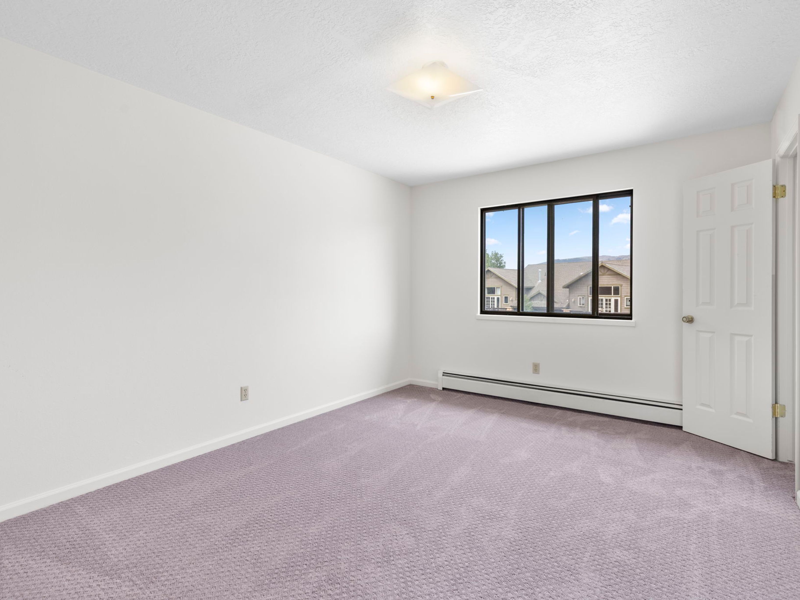This bright and neutral bedroom features light-colored walls and soft, light-purple carpeting. A large window with a black frame provides natural light and a view of the neighboring homes, while a simple white door leads out of the room. The space is illuminated by a square, flush-mount ceiling light fixture, creating a clean and versatile canvas for potential residents.