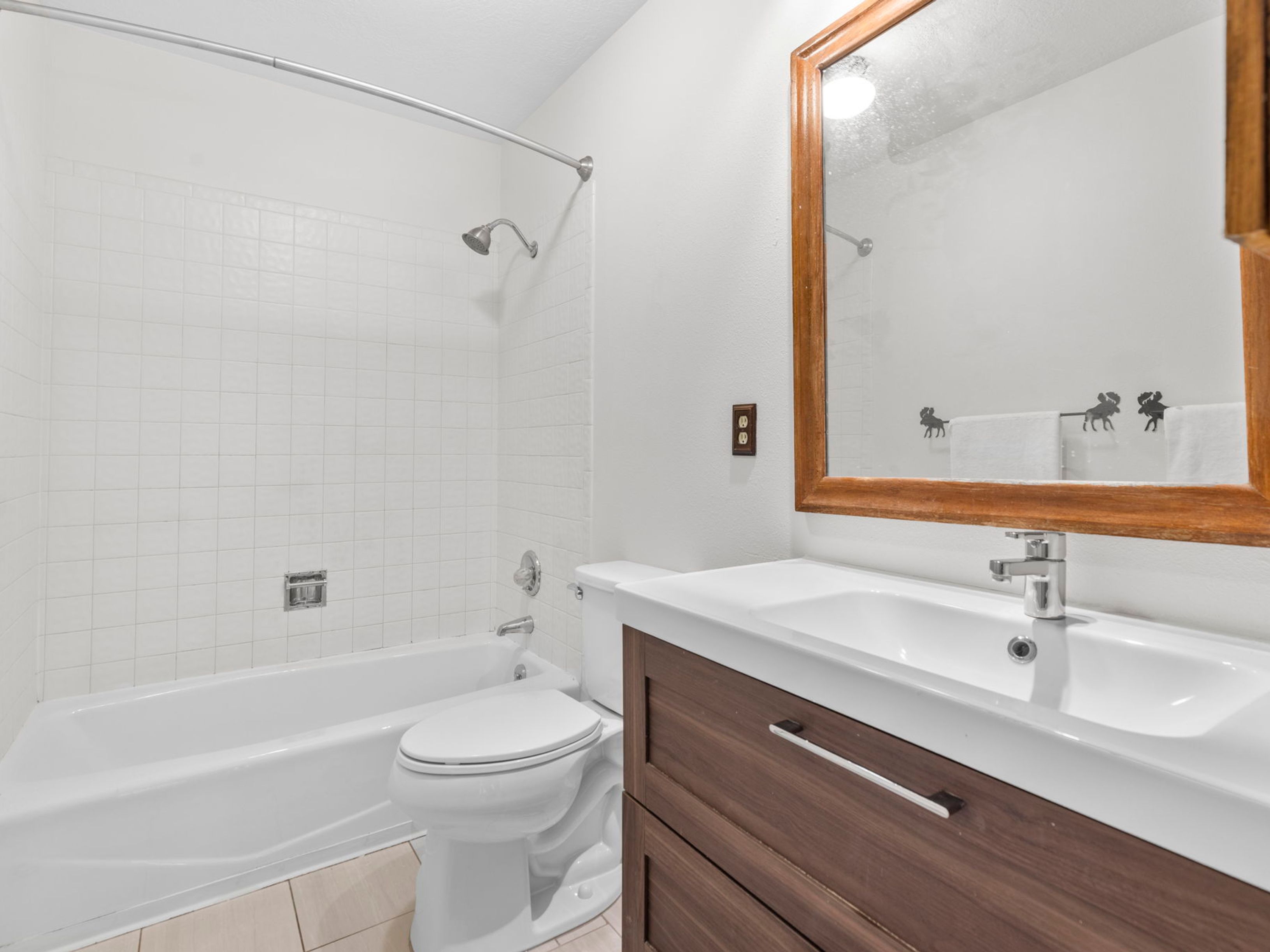 This compact bathroom features a white porcelain tub with a tiled surround and a modern vanity with a dark wood-toned cabinet. A large rectangular mirror with a warm-toned wood frame hangs above the sink, complemented by a decorative towel rack featuring moose silhouettes. The space is finished with neutral floor tiles and white walls, creating a clean and functional aesthetic.