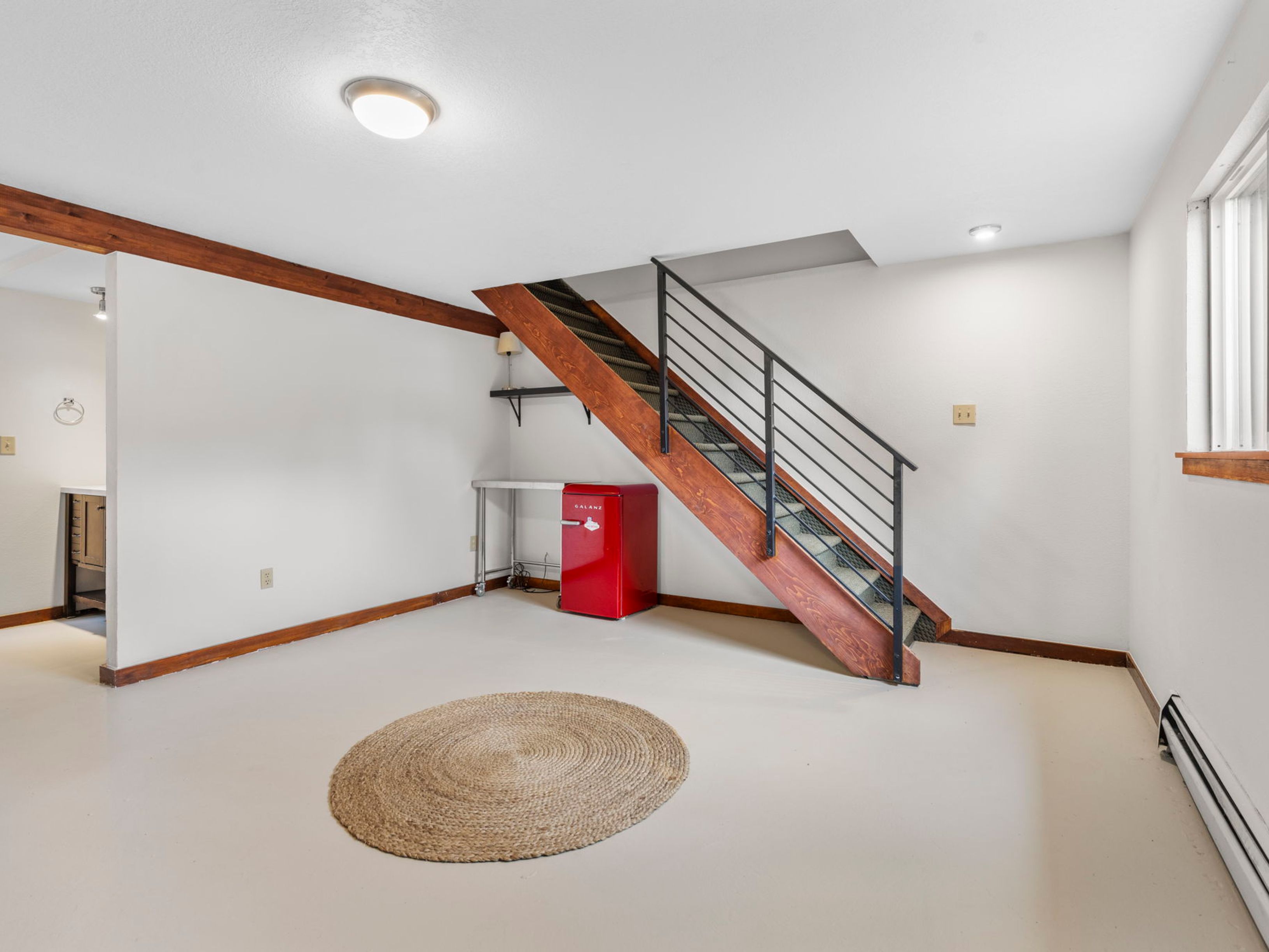 This basement space features a clean, minimalist aesthetic with light-colored flooring and white walls accented by warm wood trim. A prominent wooden staircase with a black metal railing serves as the focal point, leading to an upper level. A small red retro-style refrigerator sits beneath the stairs next to a metal utility table, while a round woven rug adds texture to the open floor area.