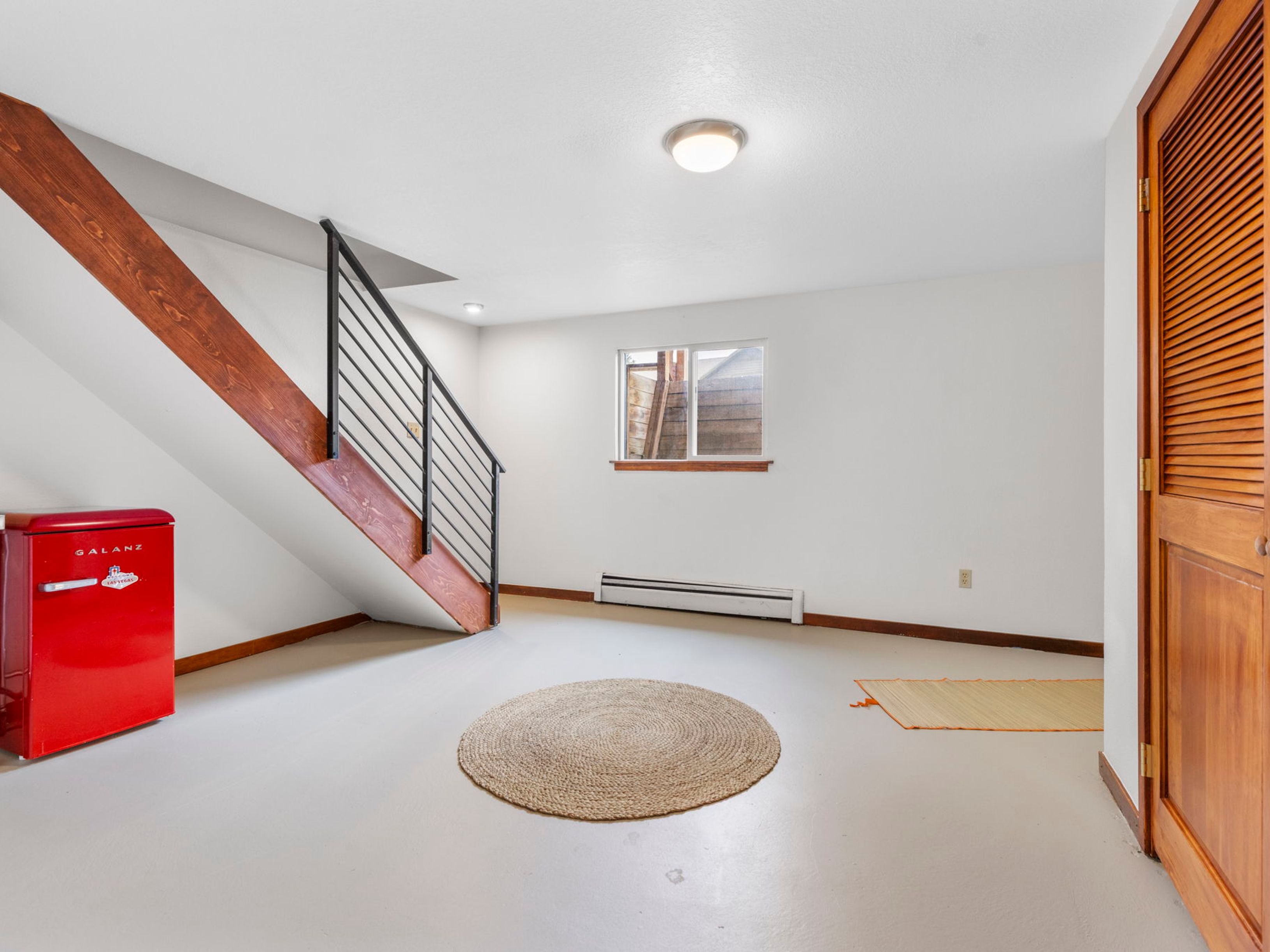 This basement space features a minimalist aesthetic with light-colored flooring and white walls, creating a bright and open atmosphere. A prominent wooden staircase with a black metal railing serves as a focal point, while a small red retro-style refrigerator adds a pop of color to the corner. A circular woven rug sits in the center of the room, and a window provides natural light, looking out toward an exterior stairwell.
