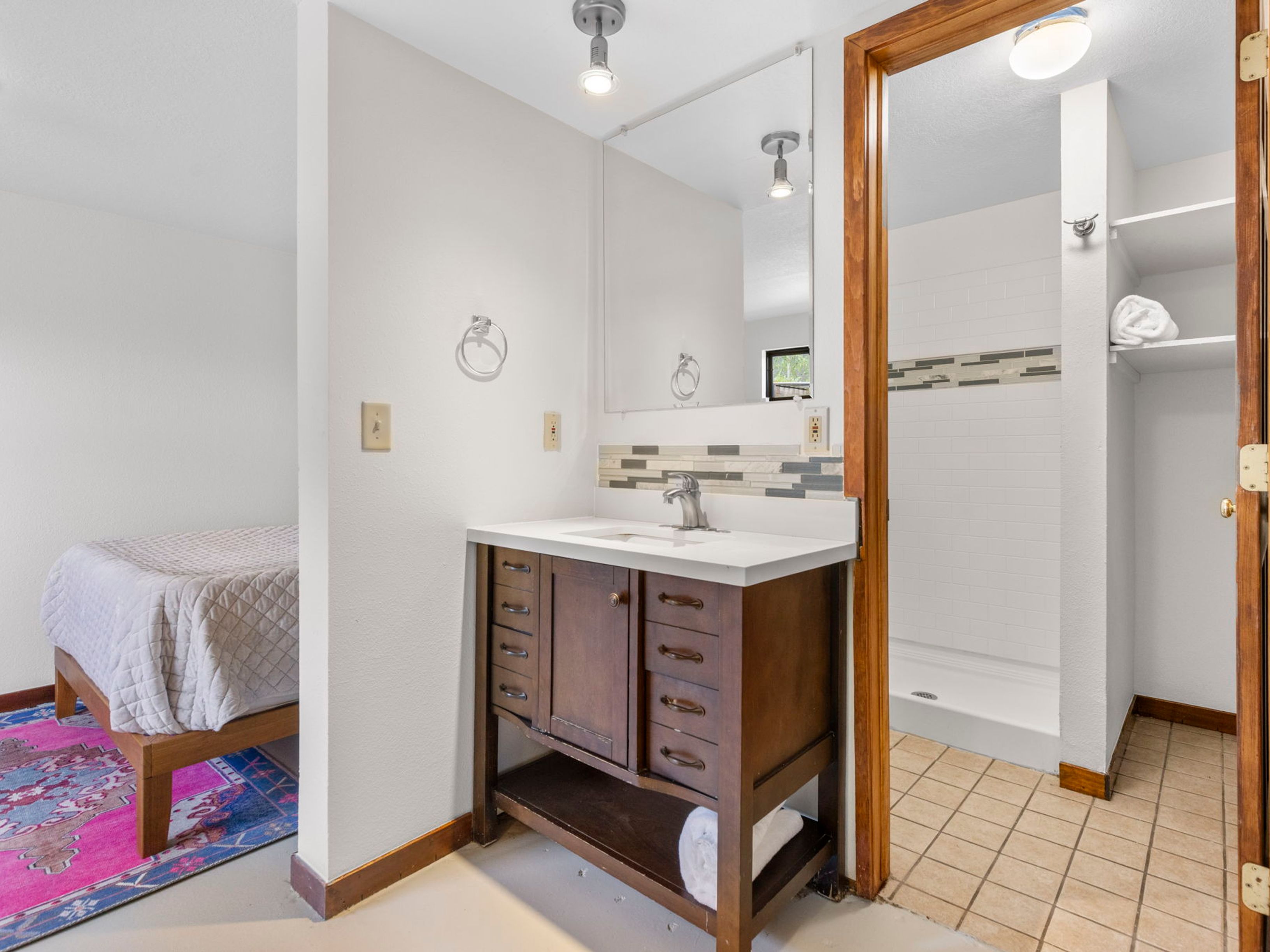 This bathroom features a dark wood vanity with a white countertop and a decorative tile backsplash, positioned adjacent to a walk-in shower area. A mirror is mounted above the vanity, and a small shelf unit is built into the shower wall for storage. The perspective captures the vanity area in the foreground with a glimpse of an adjacent bedroom space to the left, creating an open-concept feel.