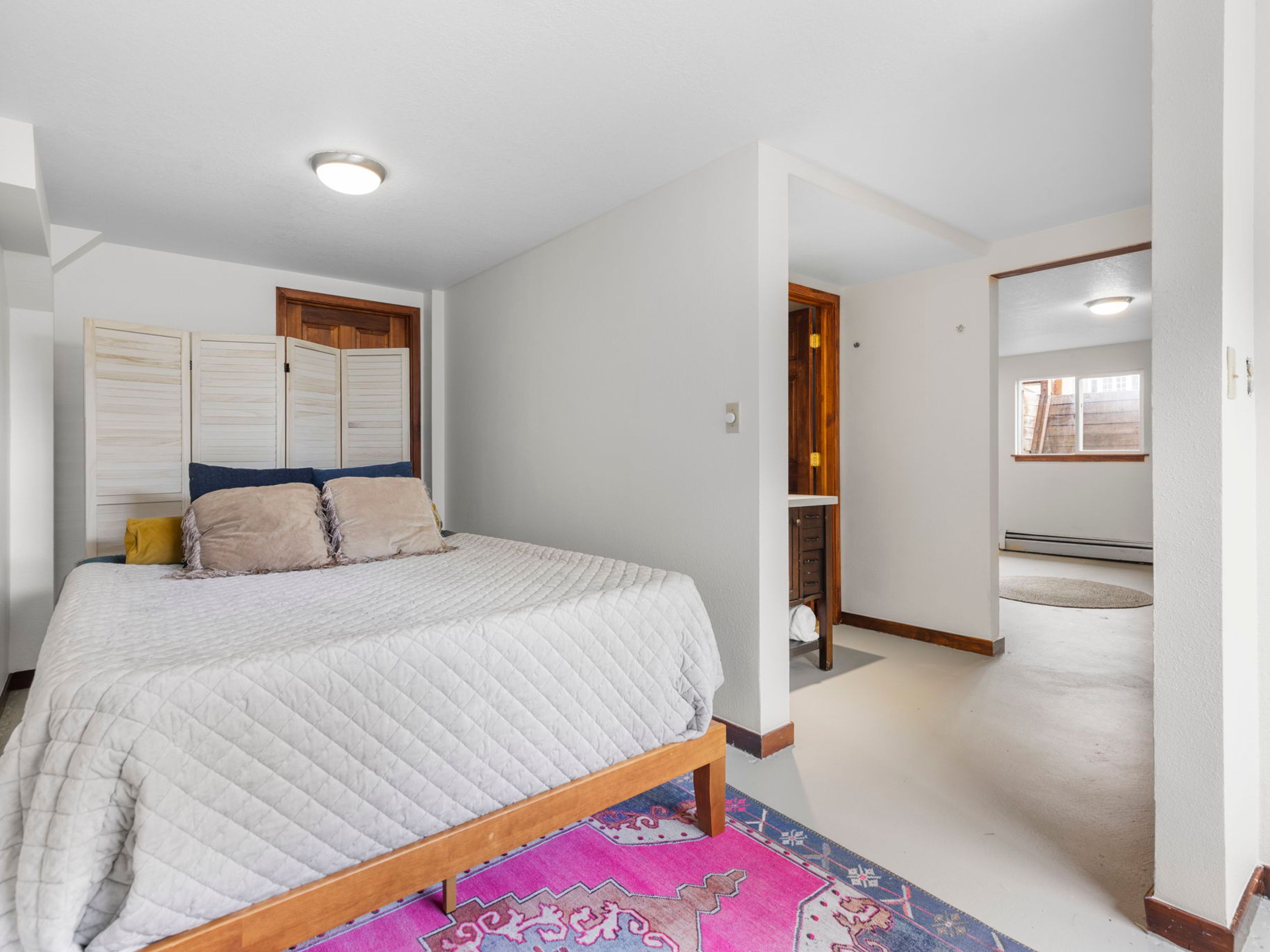 This bedroom features a light-colored bed frame with a quilted white comforter and a decorative folding screen acting as a headboard. A vibrant, patterned pink and blue rug adds a pop of color to the neutral flooring, while an open doorway leads to an adjacent room with a window. The space has a simple, functional layout with a small vanity area visible near the doorway.