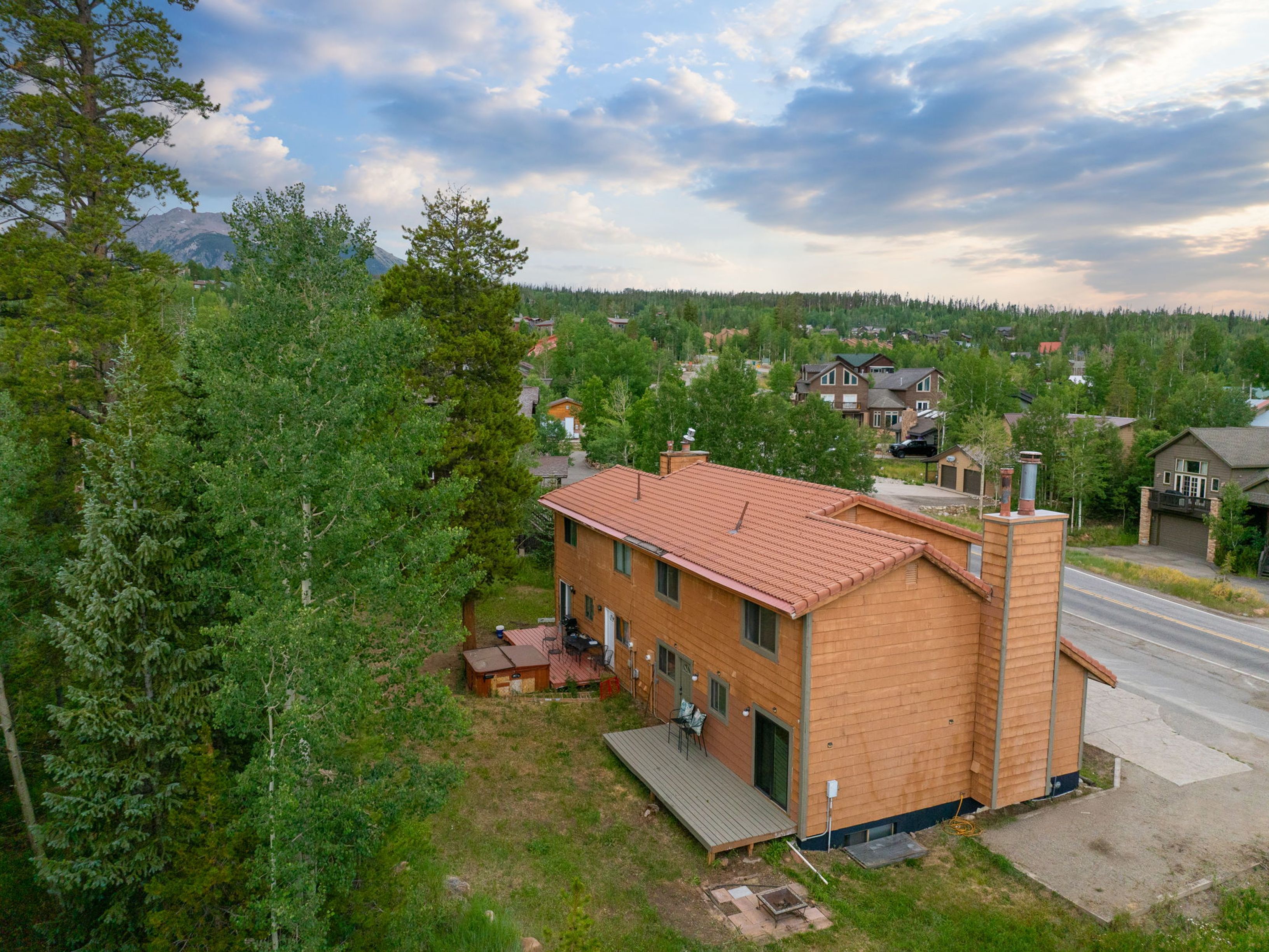 This aerial perspective captures a two-story home with a distinctive red-tiled roof, situated in a scenic, wooded mountain setting. The property features a rear deck, a small patio area with a fire pit, and a hot tub, all nestled against a backdrop of dense forest and distant peaks. A paved road runs alongside the house, providing a clear view of the surrounding residential neighborhood and natural landscape.