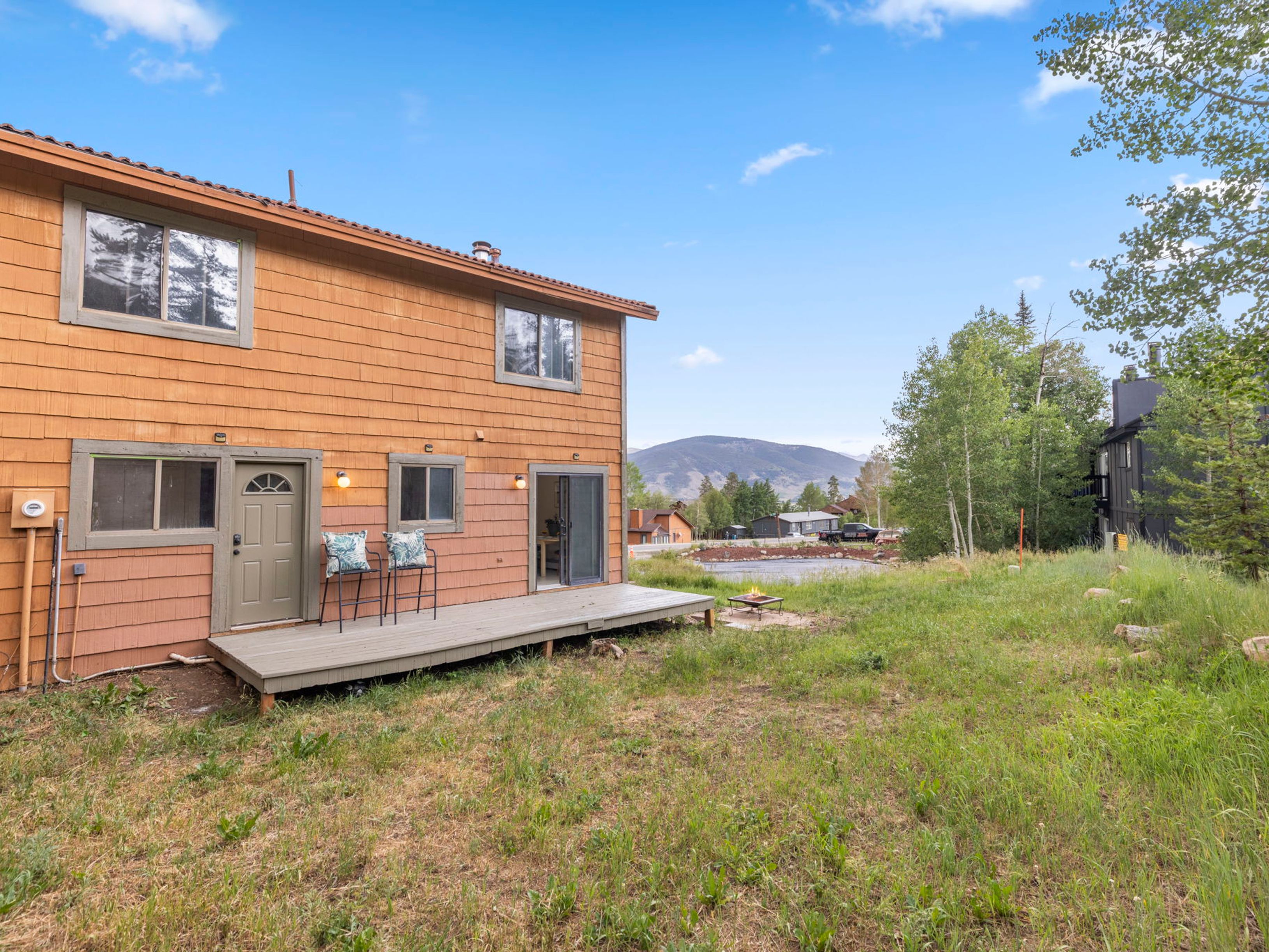 This rear view of a two-story home features warm-toned wood shingle siding and a simple, functional deck accessible via a sliding glass door. The property includes a grassy backyard that transitions into a natural, open landscape with mountain views in the distance. A small fire pit sits on the lawn, enhancing the outdoor living potential of this mountain-style residence.