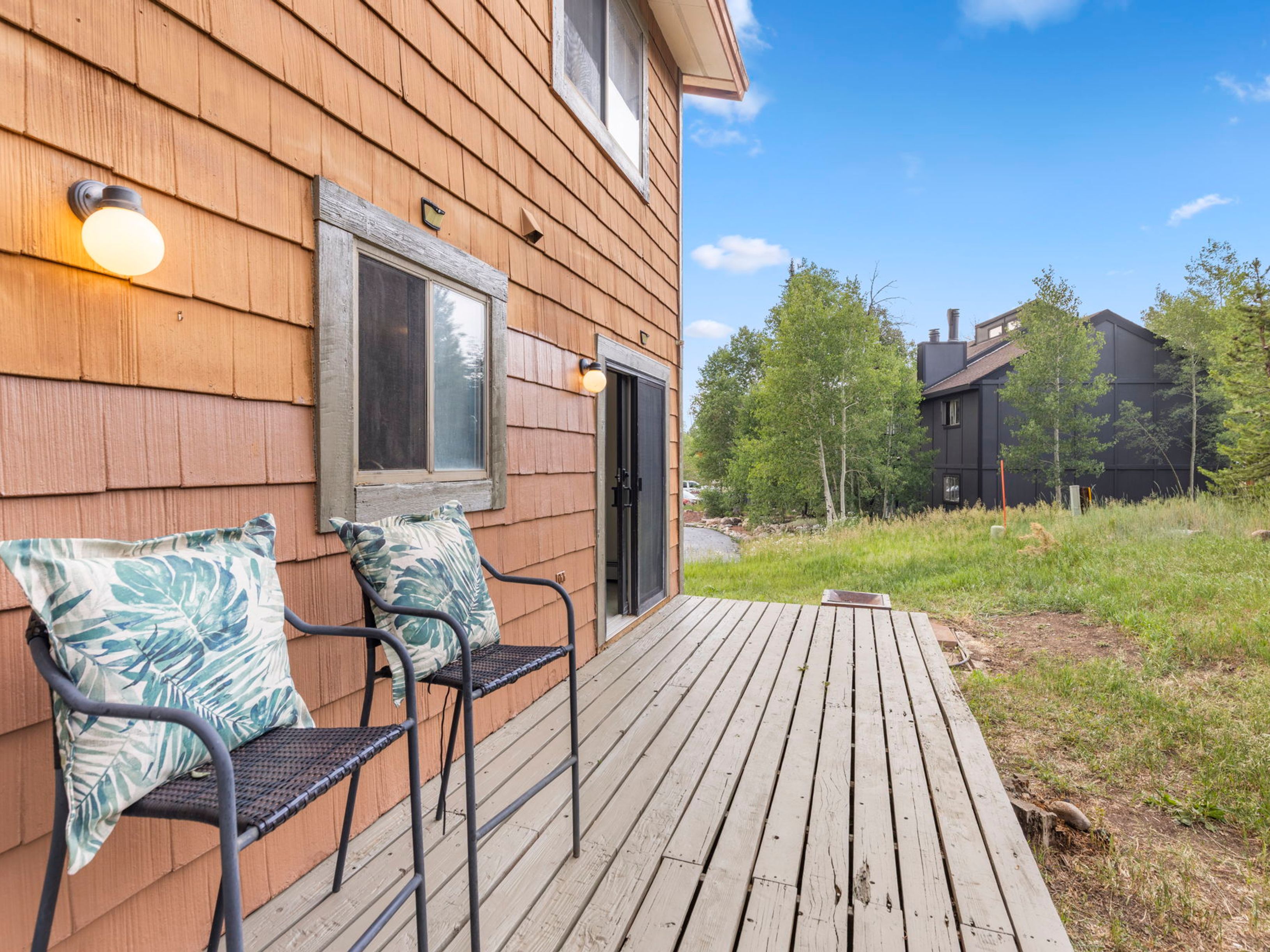 This image captures a wooden deck attached to the side of a home with cedar-shake siding, featuring two chairs with patterned cushions. The deck extends toward a grassy area and a neighboring dark-sided building, framed by mature trees and a clear blue sky. The perspective is from the deck looking outward, highlighting the outdoor living space and the surrounding natural landscape.