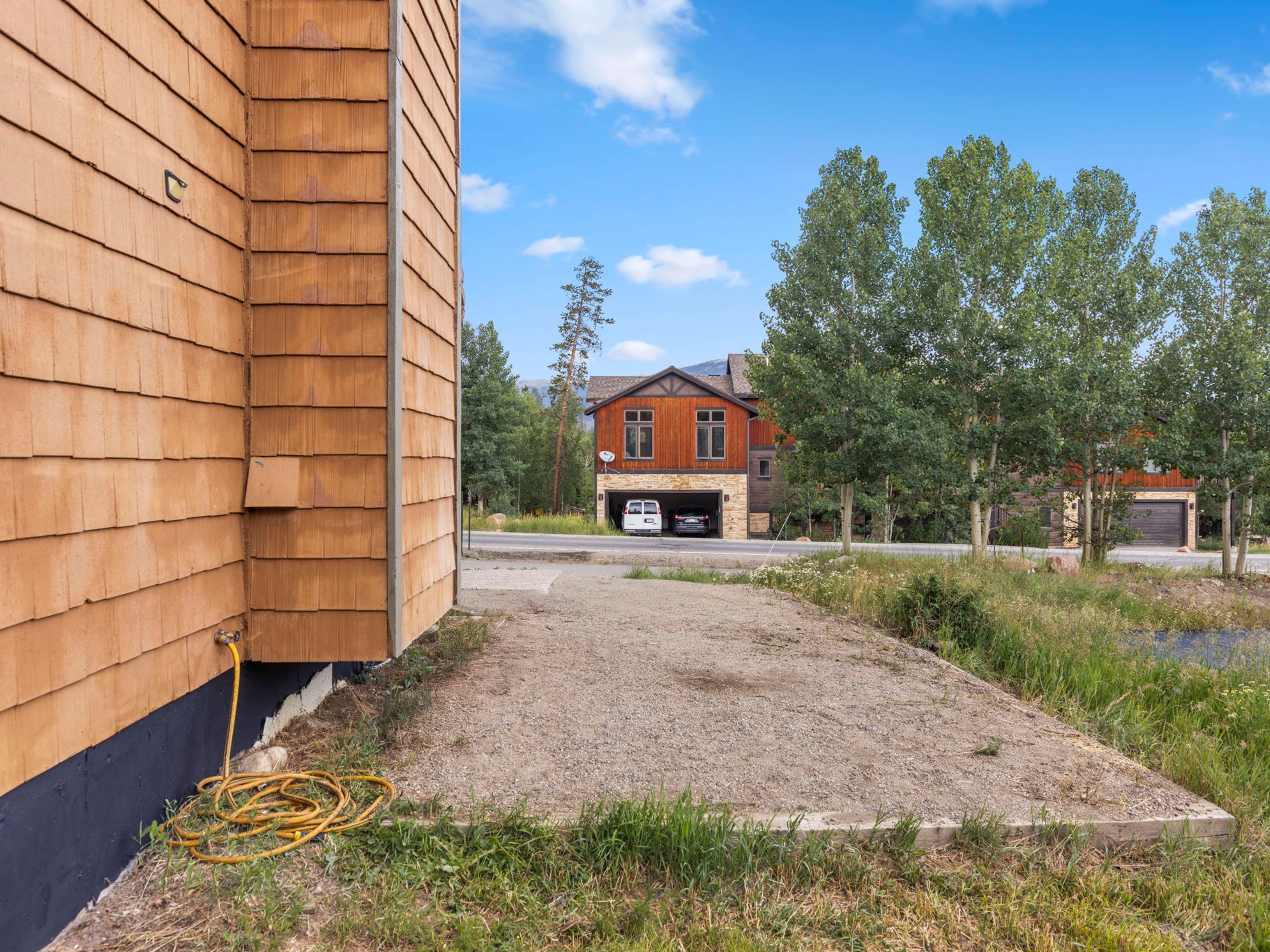 This side view captures the exterior of a home featuring warm-toned wood shingle siding and a prominent yellow garden hose coiled on the ground. A gravel-filled rectangular area sits in the foreground, bordered by wooden beams, leading toward a paved road and a neighboring multi-story house with a garage. The scene is set under a bright blue sky with scattered clouds, highlighting the surrounding trees and natural landscape.