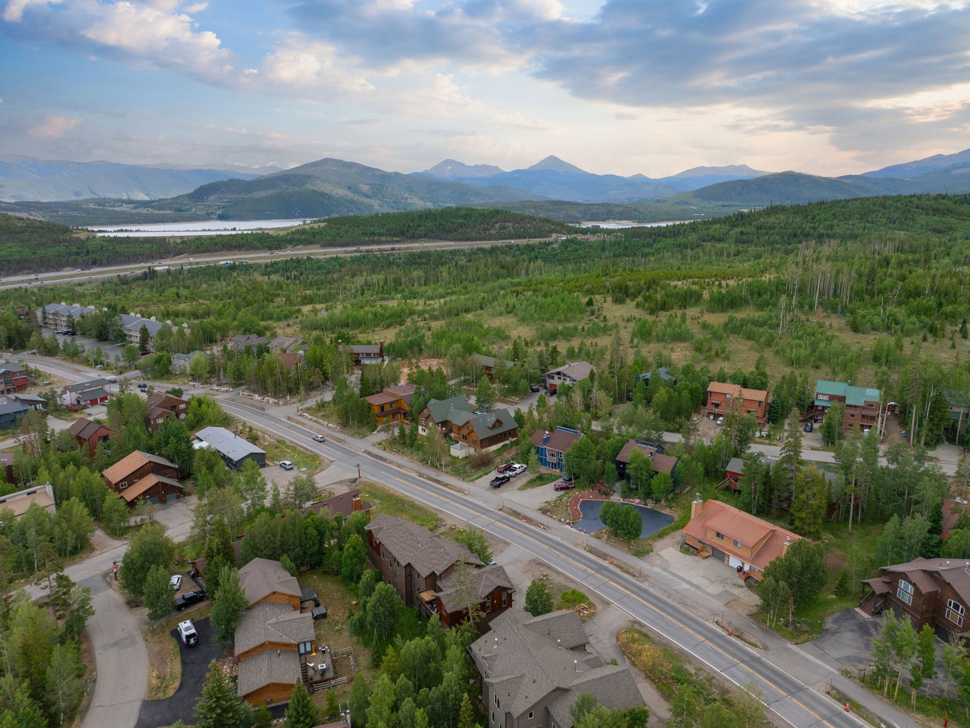 This high-angle aerial view captures a residential neighborhood nestled in a lush, forested mountain valley. The scene features a winding road flanked by various multi-story homes with diverse architectural styles, set against a backdrop of expansive green hills and distant, snow-capped mountain peaks under a dramatic, cloud-filled sky. The perspective emphasizes the community's integration with the natural landscape and its proximity to a large body of water in the distance.