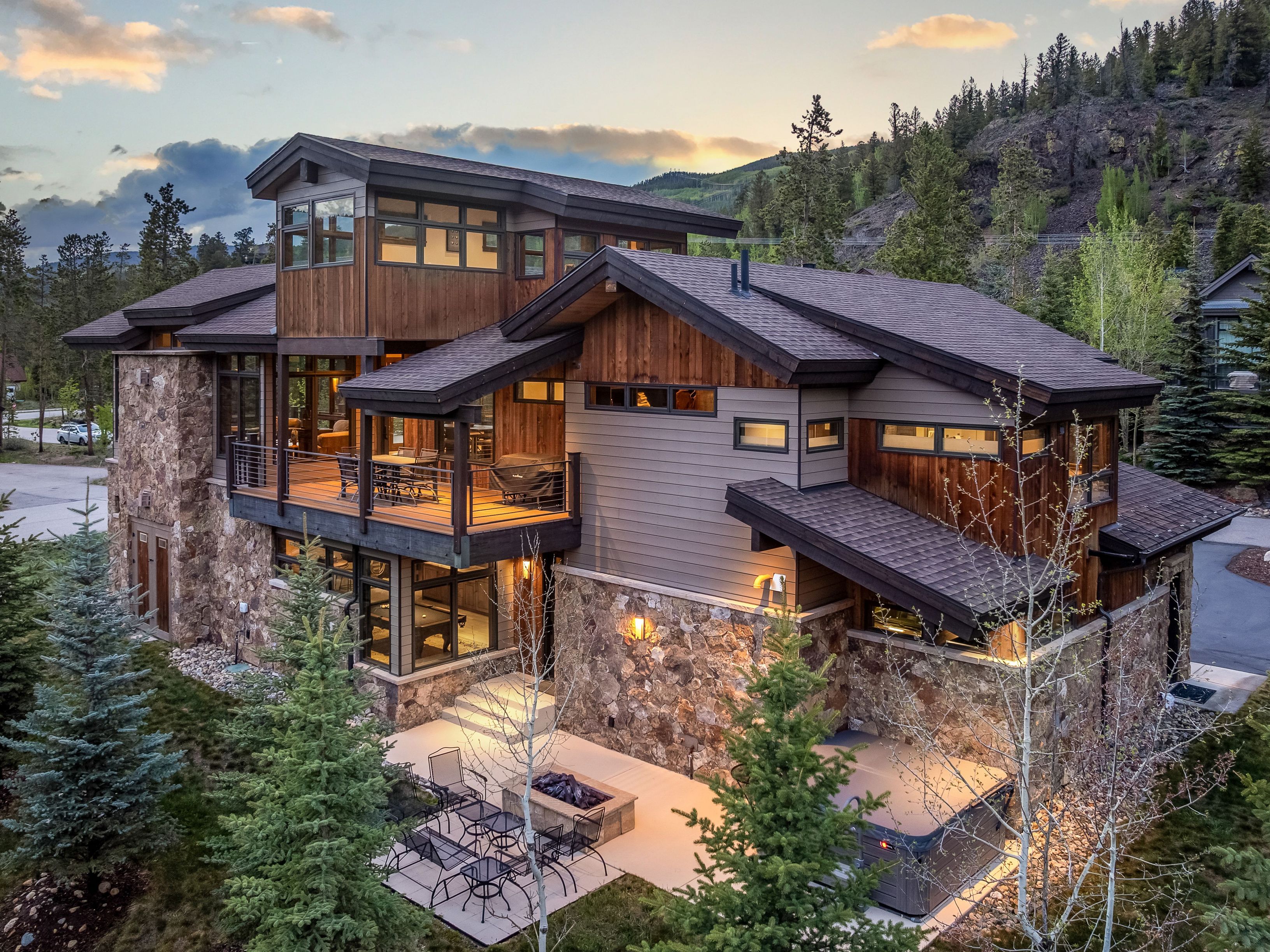 This elevated perspective captures a luxurious mountain-style residence at dusk, showcasing a sophisticated blend of stone masonry and warm-toned wood siding. The home features a multi-level design with a spacious upper-level deck and a ground-level patio area equipped with a fire pit and a hot tub. Surrounded by evergreen trees and a rugged hillside, the property exudes a serene, high-end retreat atmosphere.