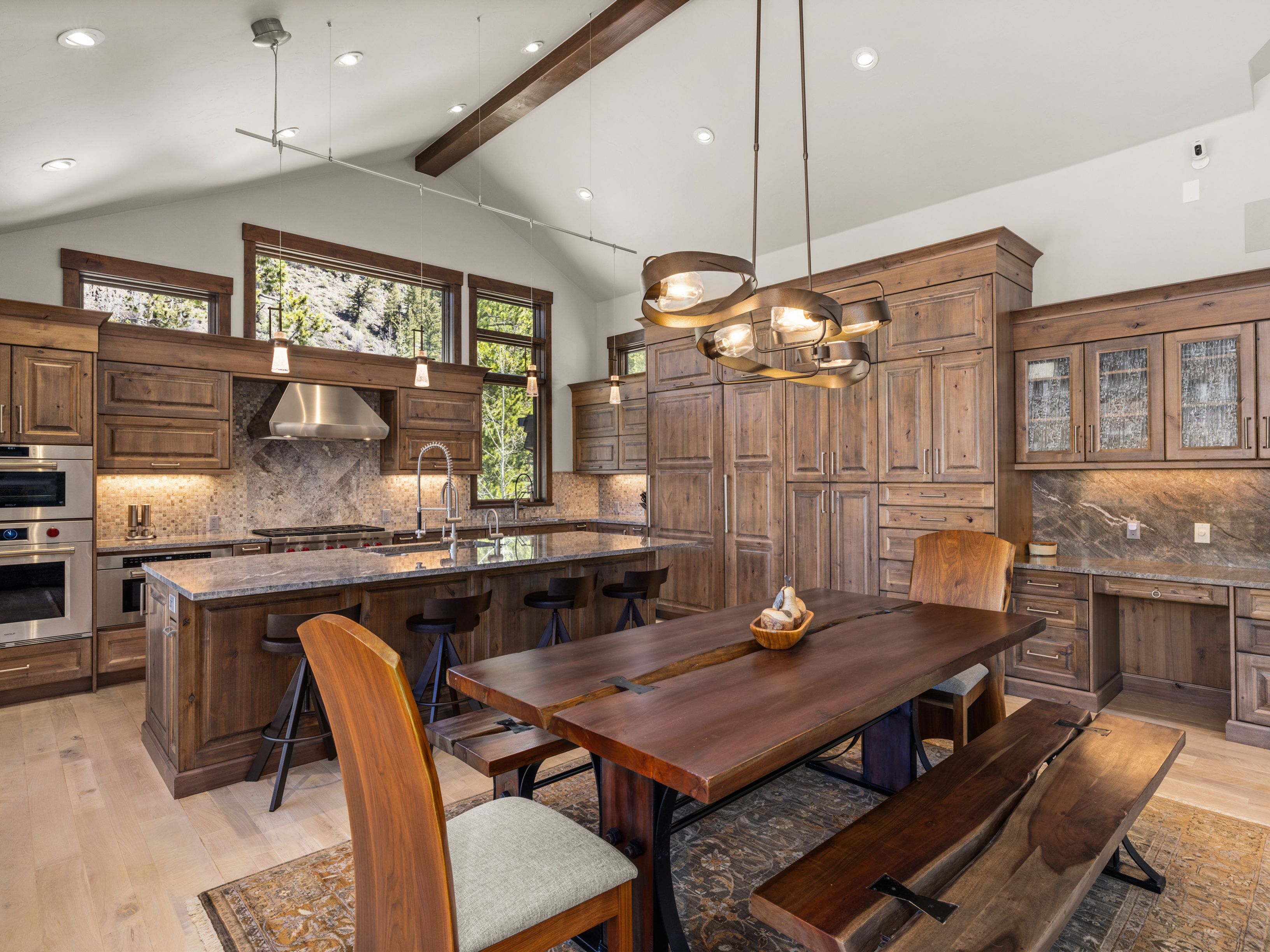 This spacious, rustic-inspired kitchen features warm wood cabinetry, a large central island with bar seating, and a dedicated dining area with a live-edge wood table. The room is illuminated by a unique, multi-ring chandelier and recessed lighting, while large windows provide natural light and views of the outdoors. The space combines high-end stainless steel appliances with natural textures, creating a sophisticated yet cozy atmosphere.