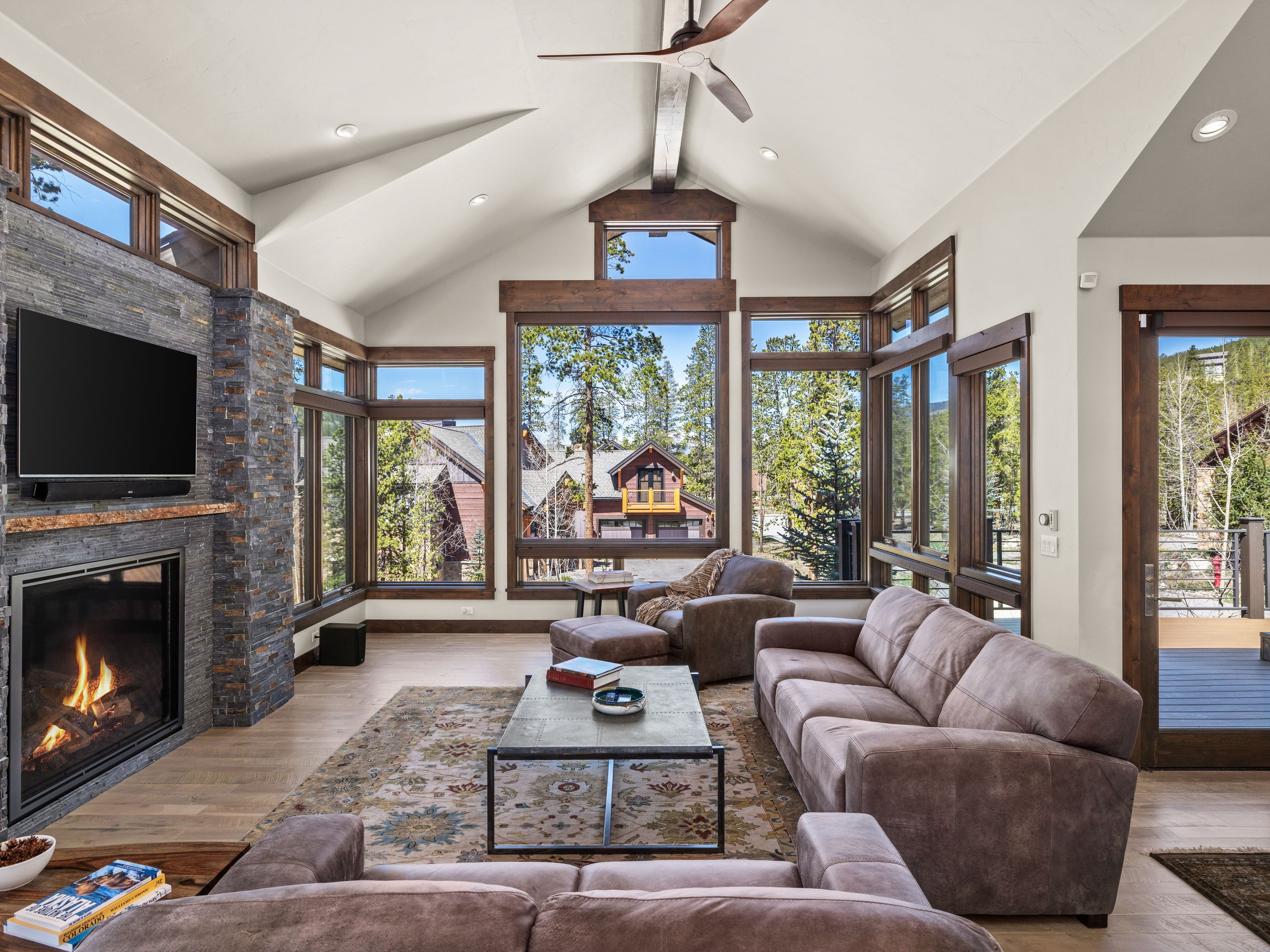 This spacious living room features a vaulted ceiling with exposed beams and a large stone-clad chimney breast housing a fireplace. The room is furnished with comfortable brown leather seating and a central metal-framed coffee table, all arranged on a patterned area rug. Large windows provide expansive views of the surrounding pine forest and neighboring homes, creating a bright and inviting mountain-style atmosphere.