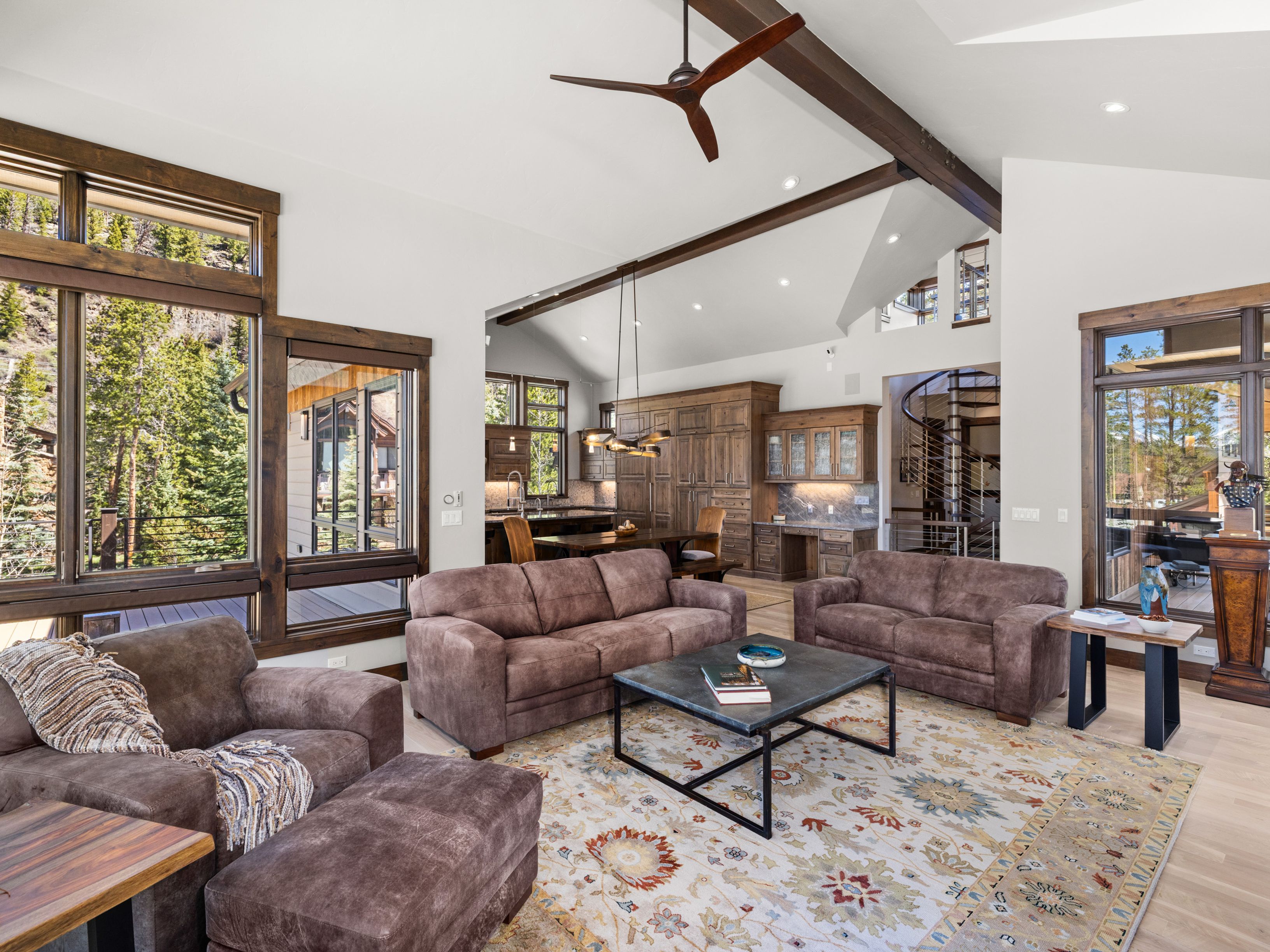 This expansive open-concept living area features vaulted ceilings with exposed dark wood beams and a large ceiling fan. The space is anchored by a patterned area rug, a dark metal-framed coffee table, and plush brown seating, with a kitchen and dining area visible in the background. Large windows provide abundant natural light and views of the surrounding forest, while a spiral staircase adds a unique architectural element to the home's interior.