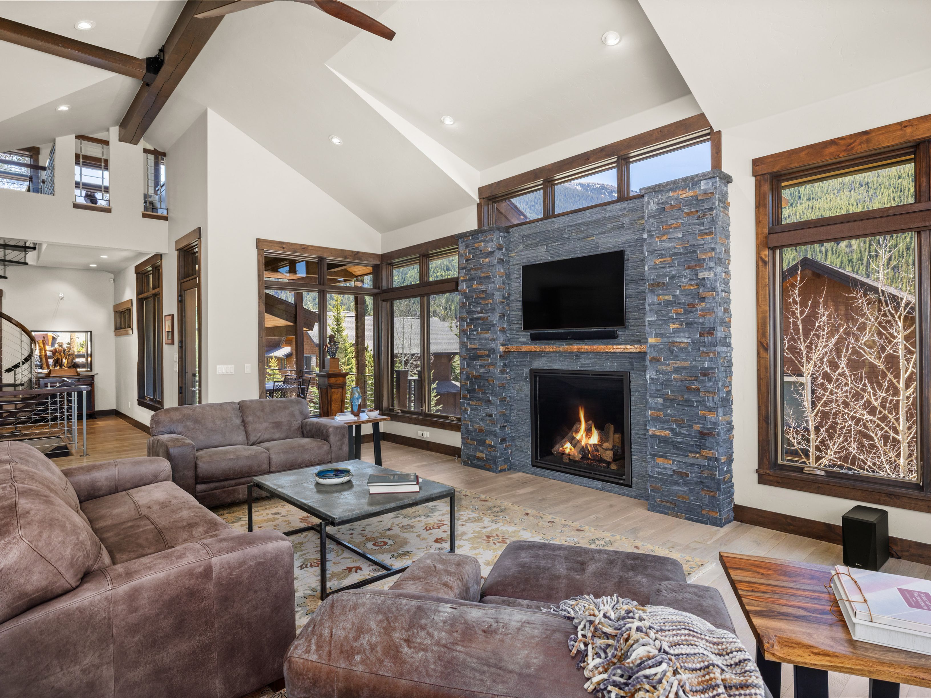 This spacious living room features a vaulted ceiling with exposed wooden beams and a prominent stone-clad chimney breast housing a fireplace and television. Large windows with dark wood trim frame mountain views, while plush brown sofas and a patterned area rug create a cozy, inviting atmosphere. The open-concept layout flows into a staircase area, emphasizing a rustic yet refined mountain lodge aesthetic.