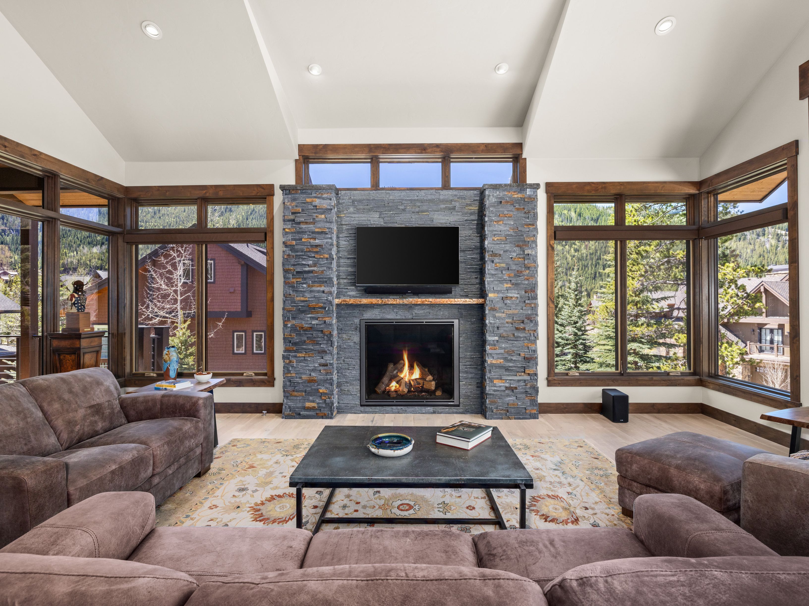 This spacious living room features a prominent stone-clad chimney breast housing a fireplace and a mounted television, serving as the central focal point. The room is framed by large windows on either side that offer expansive views of the surrounding mountain landscape, while a plush, dark-toned sectional sofa and a central coffee table create a comfortable seating area. The vaulted ceiling and light-toned wood flooring contribute to an airy, mountain-lodge aesthetic that feels both luxurious and inviting.