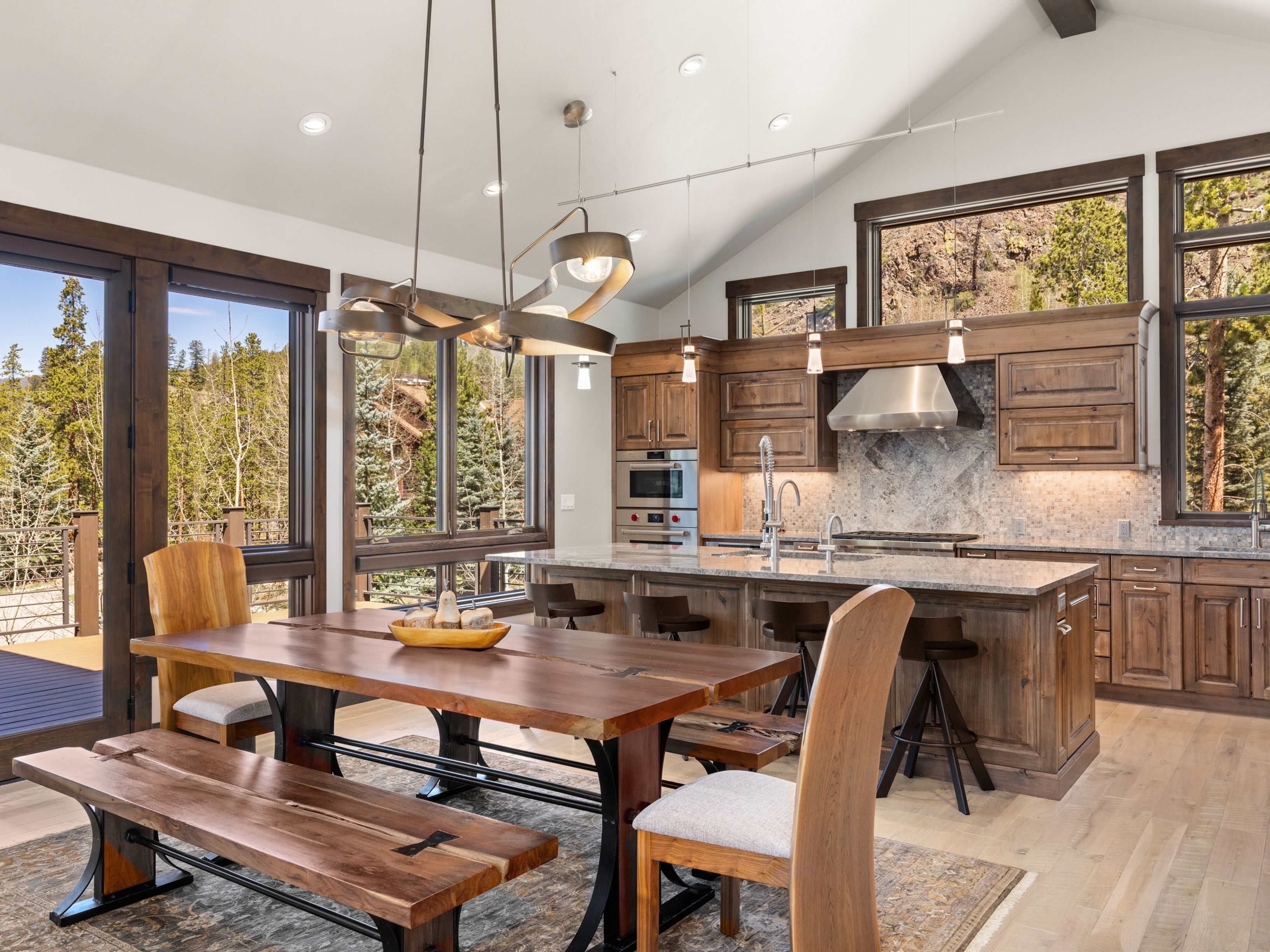 This open-concept space features a rustic-modern kitchen with a large central island, warm wood cabinetry, and stainless steel appliances. A substantial dining table with a live-edge wood top and matching bench seating sits in the foreground, creating a cohesive transition between the dining and cooking areas. Large windows provide expansive views of the surrounding forest, while a unique, sculptural chandelier hangs above the dining table to anchor the room.