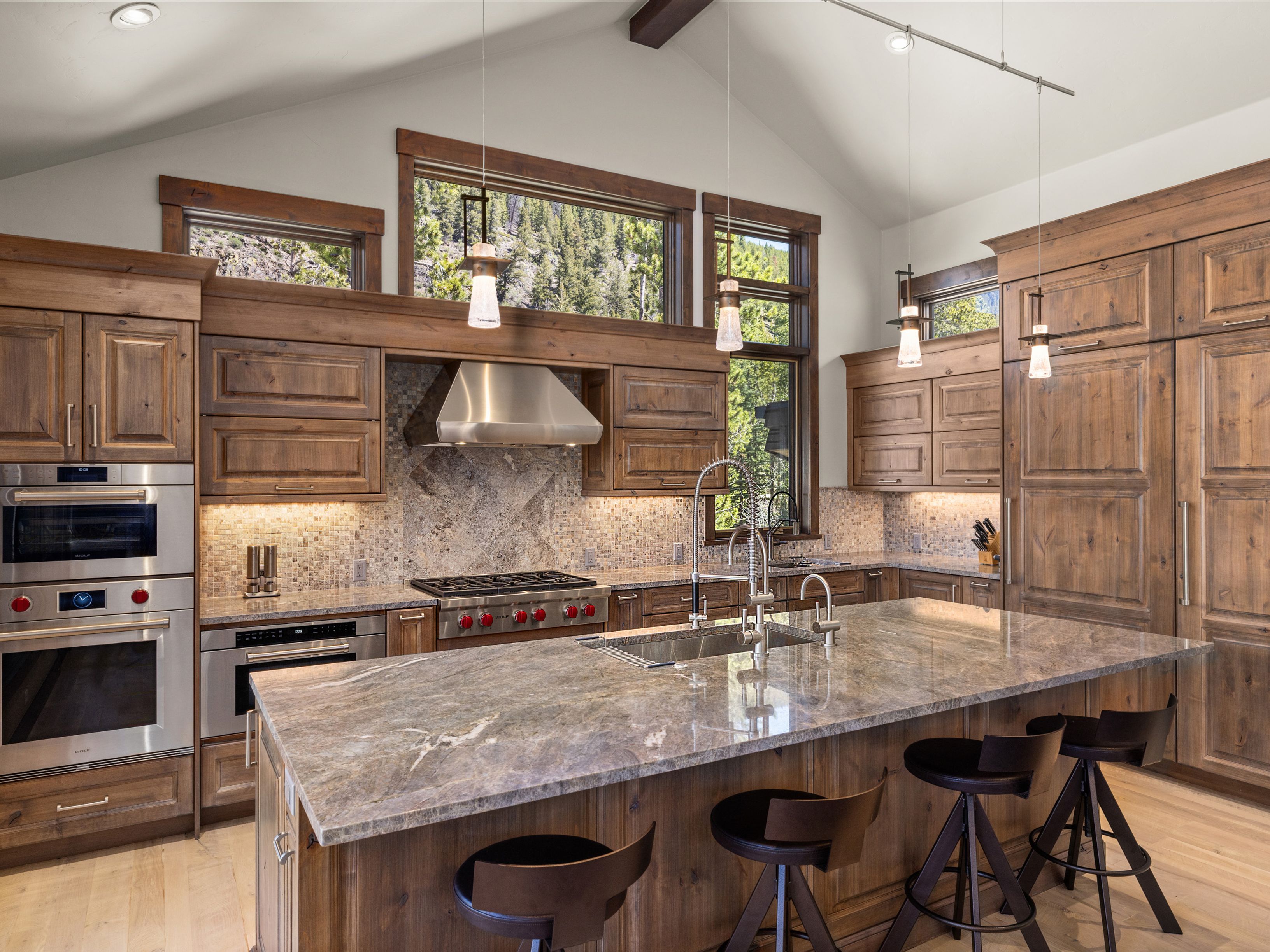 This spacious, rustic-inspired kitchen features warm-toned wood cabinetry, a large central island with stone countertops, and high-end stainless steel appliances. The room is illuminated by pendant lights and natural light streaming through large windows that offer views of the surrounding forest. The design is anchored by a stone-tiled backsplash and a professional-grade range, creating a sophisticated yet cozy atmosphere.
