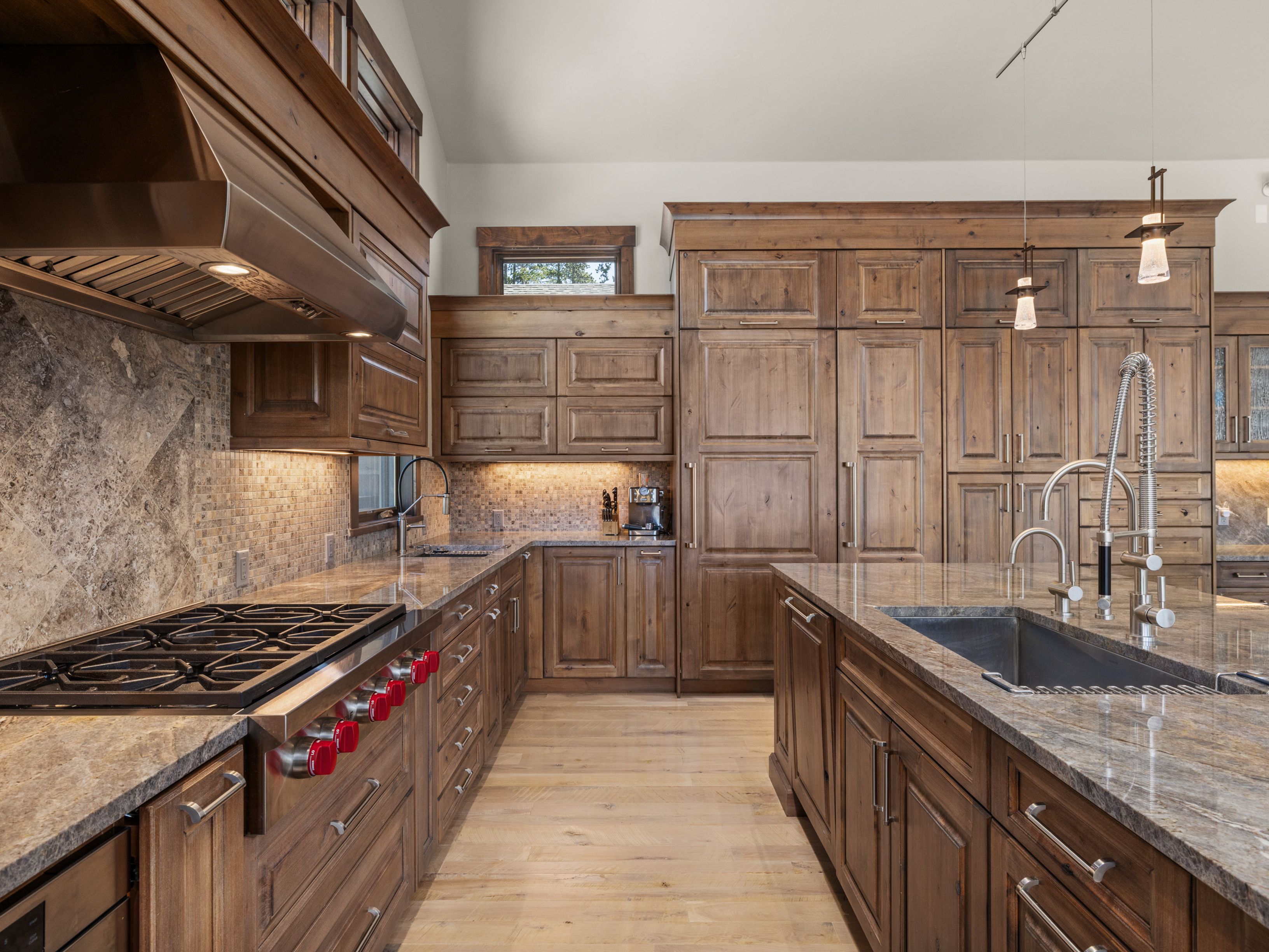 This rustic-inspired kitchen features extensive medium-toned wood cabinetry and a large central island topped with patterned stone countertops. A professional-grade range with distinctive red knobs sits beneath a large stainless steel hood, complemented by a tiled backsplash and a deep undermount sink. The space is illuminated by warm under-cabinet lighting and hanging pendant lights, creating a cozy and functional atmosphere.