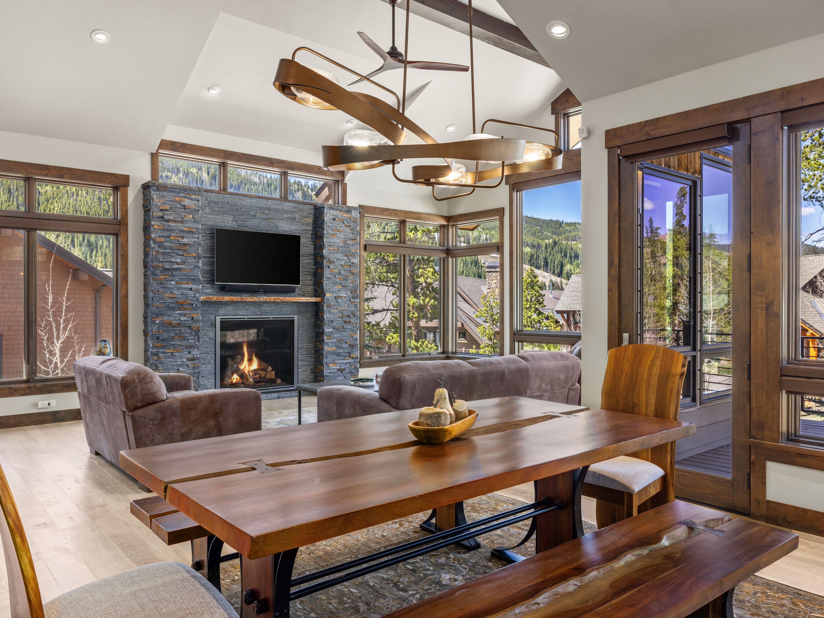 This open-concept living and dining area features a rustic aesthetic with a prominent stacked-stone chimney breast housing a fireplace and television. A substantial live-edge wood dining table serves as the focal point in the foreground, complemented by a unique, sculptural metal chandelier overhead. Large windows throughout the space provide expansive views of the surrounding mountain landscape, creating a bright and inviting atmosphere.