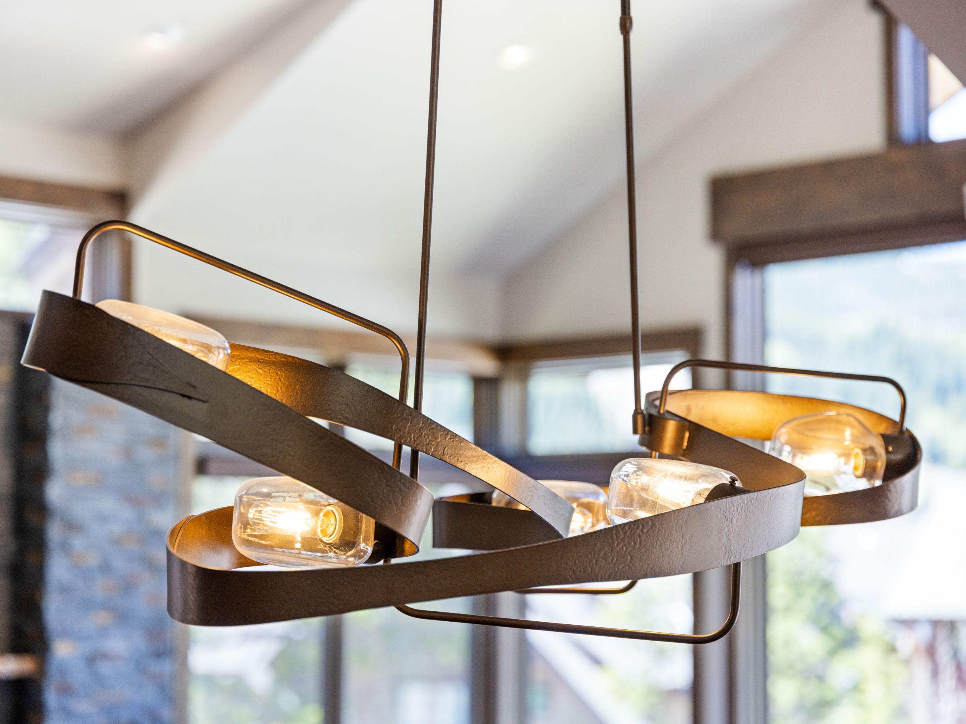 A close-up, cinematic shot focuses on a modern, sculptural chandelier featuring a dark, textured metal ribbon frame that winds around several exposed-filament glass bulbs. The warm, glowing light from the bulbs creates a cozy atmosphere, while the blurred background reveals a bright, open-concept living space with large windows and a stone-clad wall. The perspective highlights the intricate design and artistic craftsmanship of the lighting fixture as a central focal point of the room.