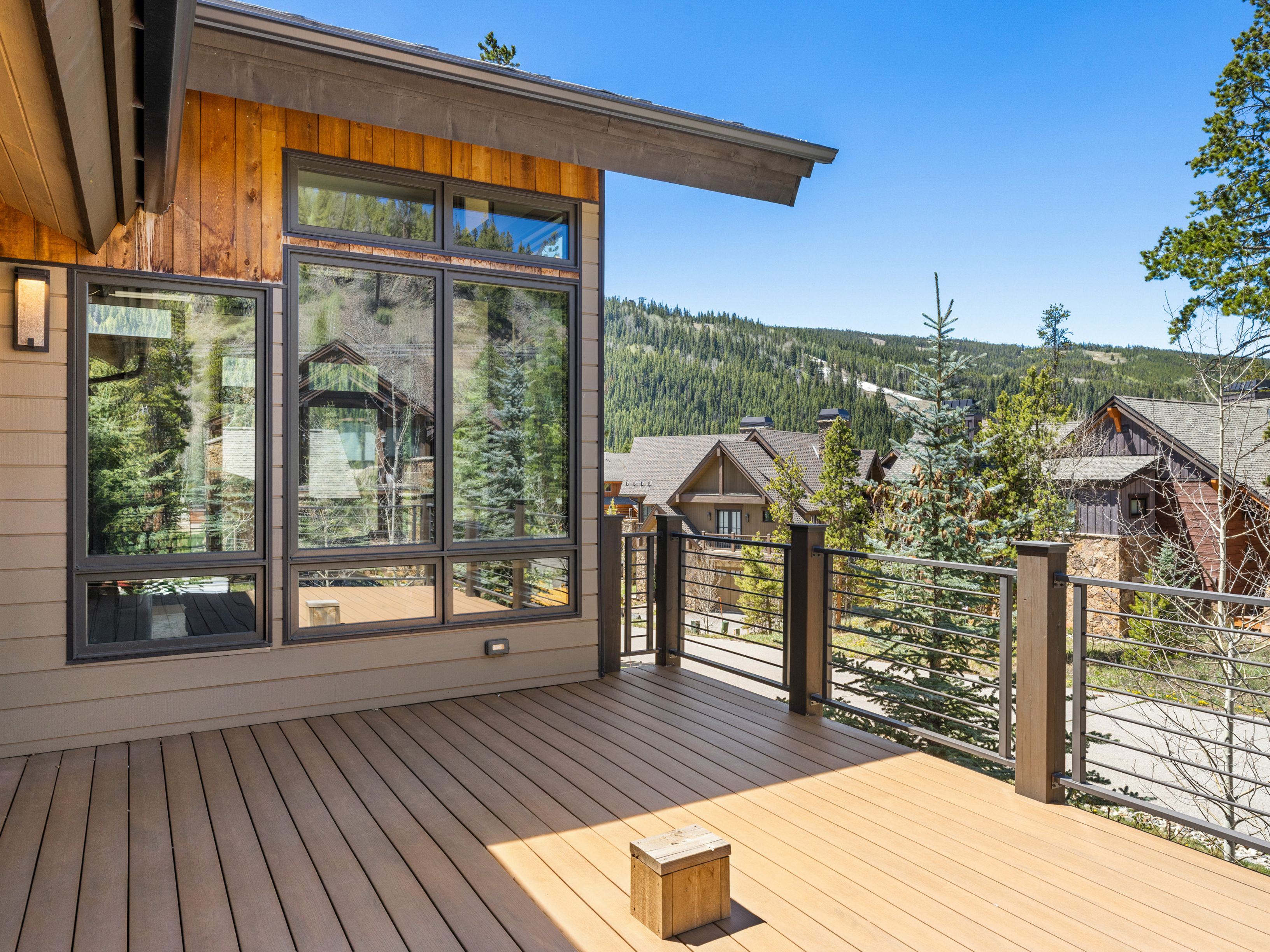 This elevated deck features composite wood-look decking and a modern metal railing system, offering expansive views of a mountainous landscape and neighboring homes. The exterior wall of the house is finished with a combination of horizontal siding and vertical wood paneling, complemented by large, dark-framed windows. The perspective captures the deck from a slightly elevated angle, emphasizing the seamless transition between indoor and outdoor living spaces.