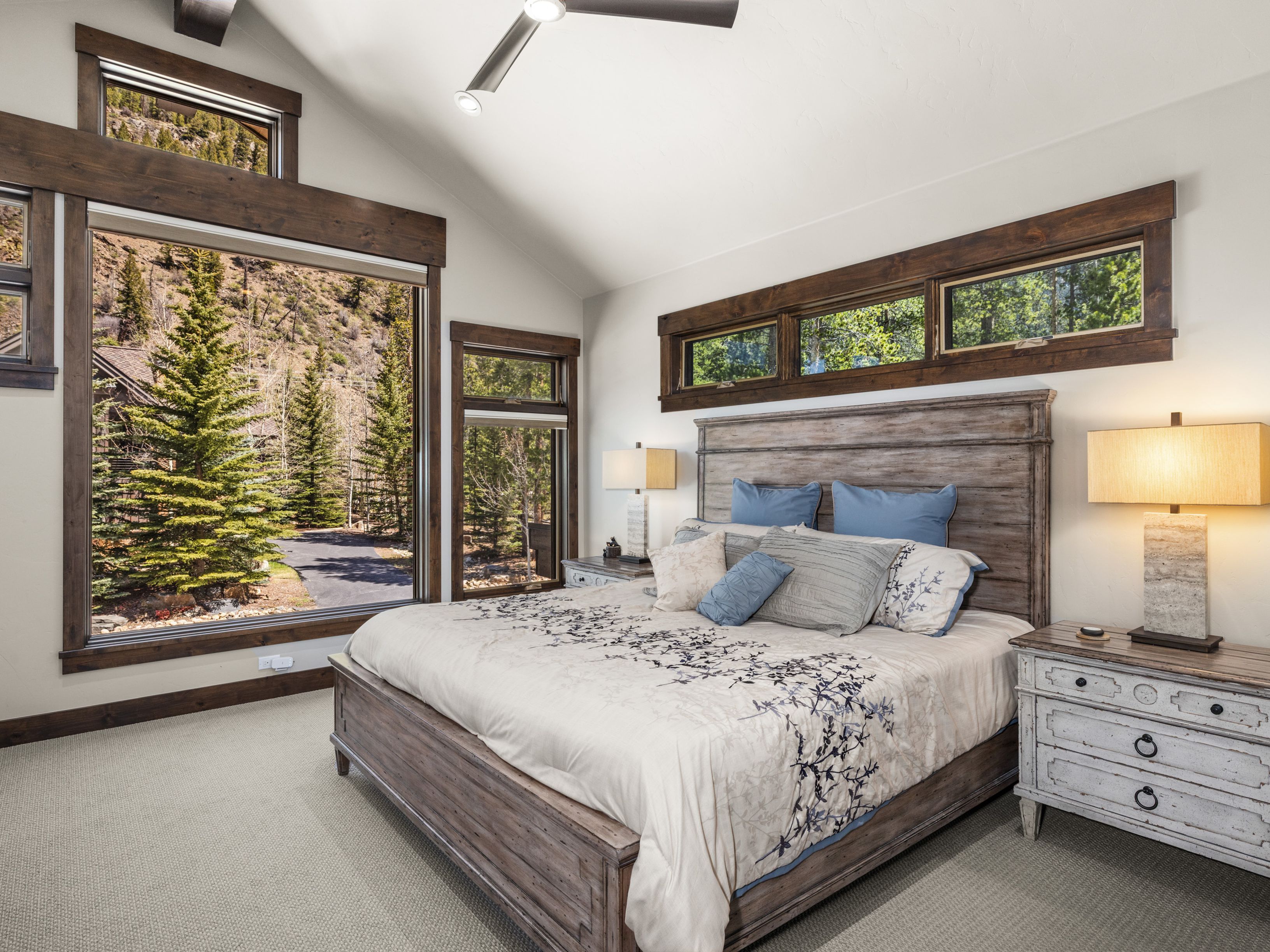 This primary bedroom features a rustic aesthetic with a large wooden bed frame and matching nightstands. The room is bathed in natural light from expansive windows that offer scenic views of the surrounding forest and hillside. A vaulted ceiling with a modern fan adds architectural interest, while the neutral color palette creates a serene and inviting atmosphere.
