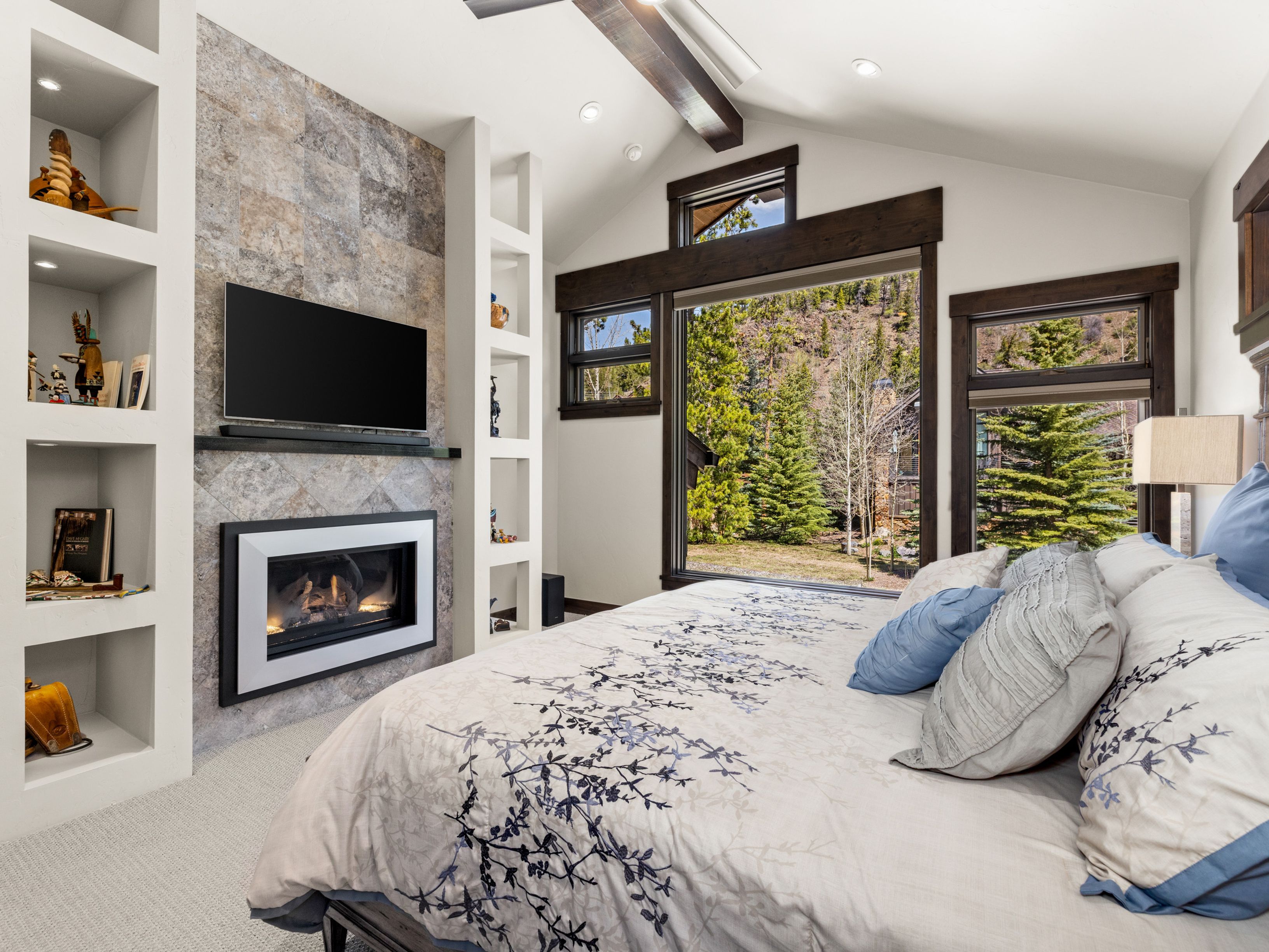 This primary bedroom features a cozy, rustic aesthetic with a large bed dressed in patterned linens and a stone-clad chimney breast housing a fireplace and television. Built-in shelving units flank the fireplace, providing display space for decor, while large windows offer a scenic view of the surrounding forest. The room is accented by dark wood trim and a vaulted ceiling, creating a warm and inviting atmosphere.