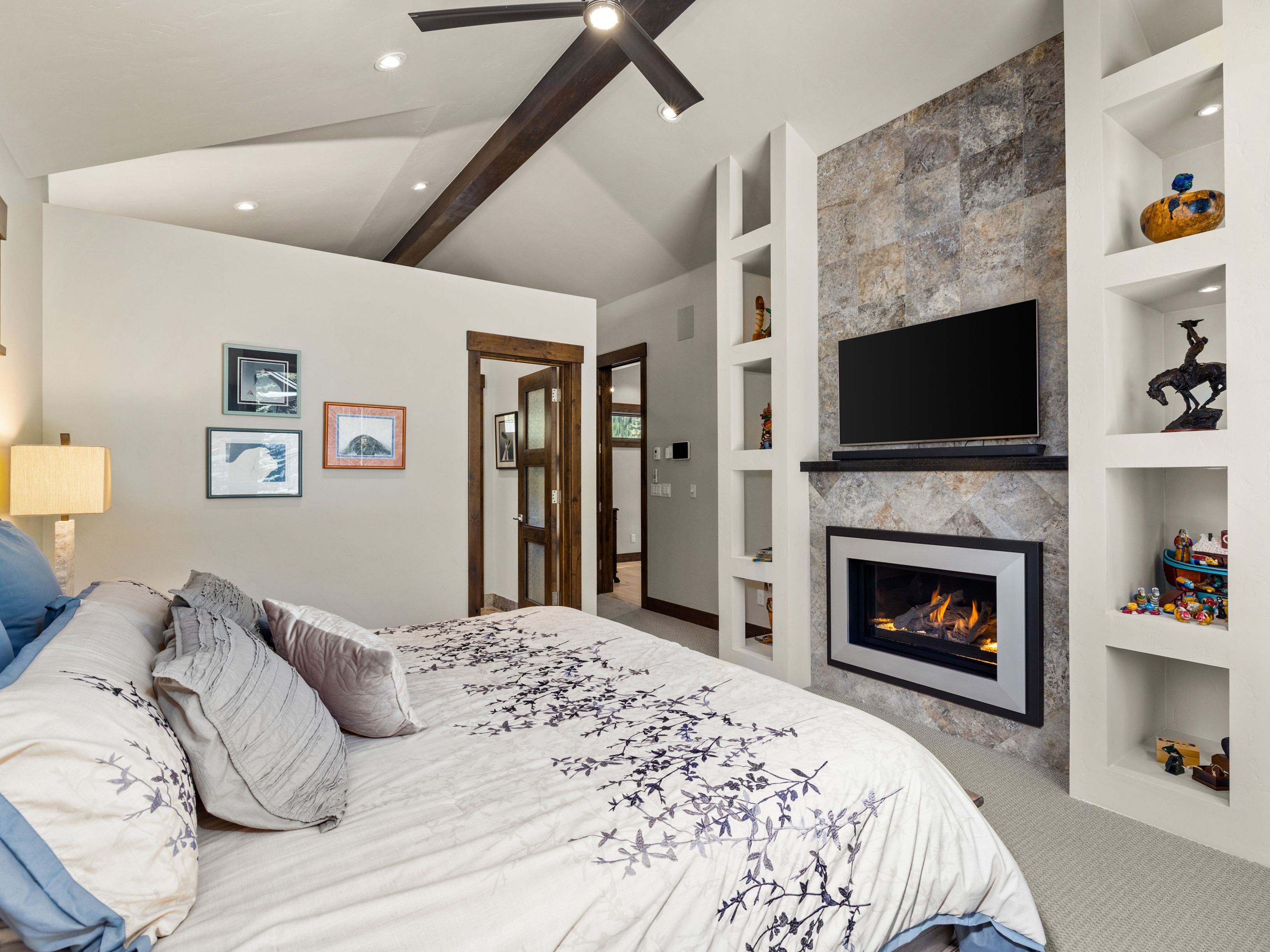 This primary bedroom features a vaulted ceiling with exposed dark wood beams and a central ceiling fan. A prominent stone-clad chimney breast serves as the focal point, housing a built-in fireplace and a wall-mounted television, flanked by custom white shelving units. The room is decorated with a patterned bedspread and framed wall art, creating a cozy and inviting atmosphere.
