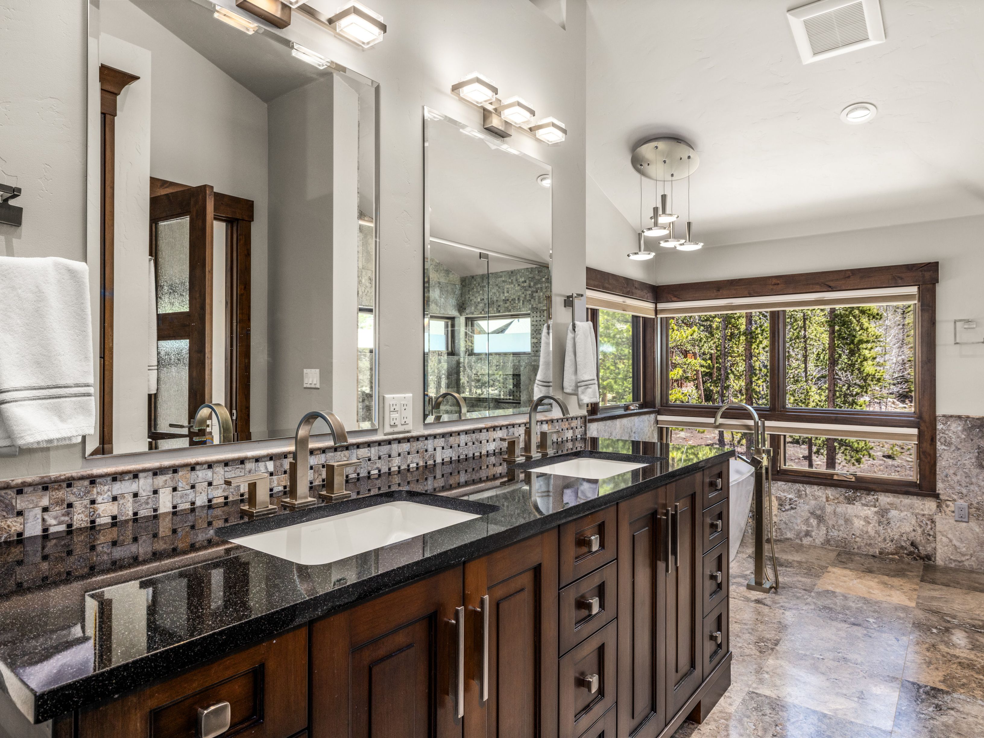 This primary bathroom features a double vanity with dark wood cabinetry, a dark stone countertop, and dual rectangular undermount sinks. Large mirrors are mounted above the vanity, complemented by modern light fixtures and a mosaic tile backsplash. The space includes a freestanding tub positioned near large windows that offer a scenic view of the surrounding forest, creating a serene and luxurious atmosphere.