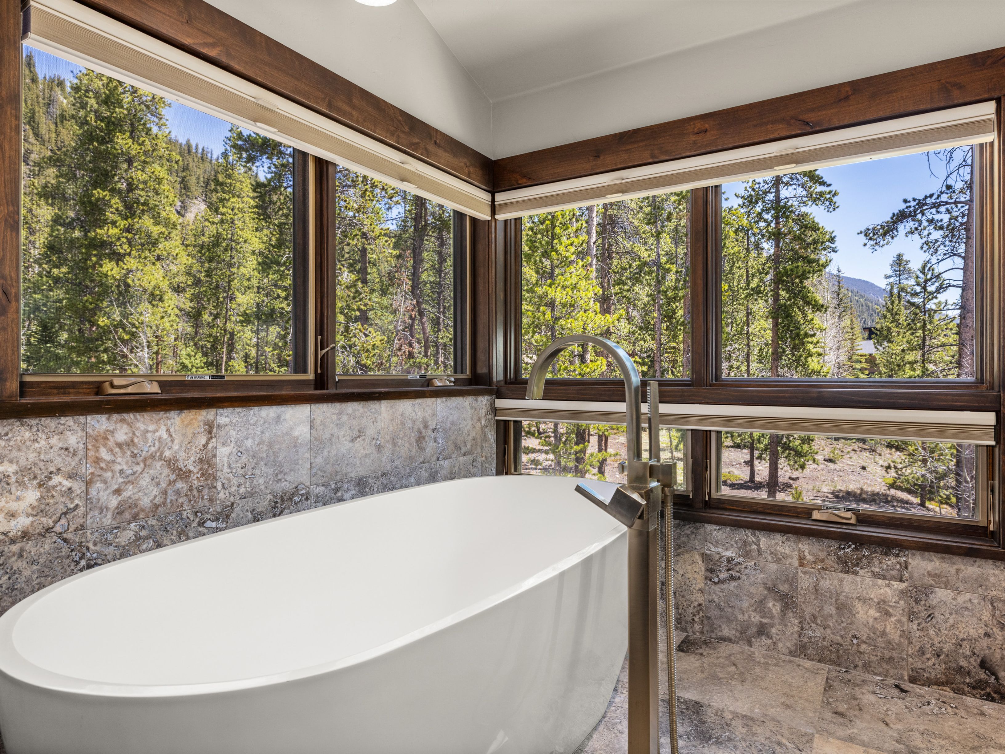 This primary bathroom features a sleek, white freestanding soaking tub positioned as the focal point against a backdrop of large, light-toned stone wall tiles. Expansive windows wrap around the corner, offering an immersive, unobstructed view of the surrounding forest and mountain landscape. A modern, metallic floor-mounted faucet adds a contemporary touch to the serene, nature-inspired space.