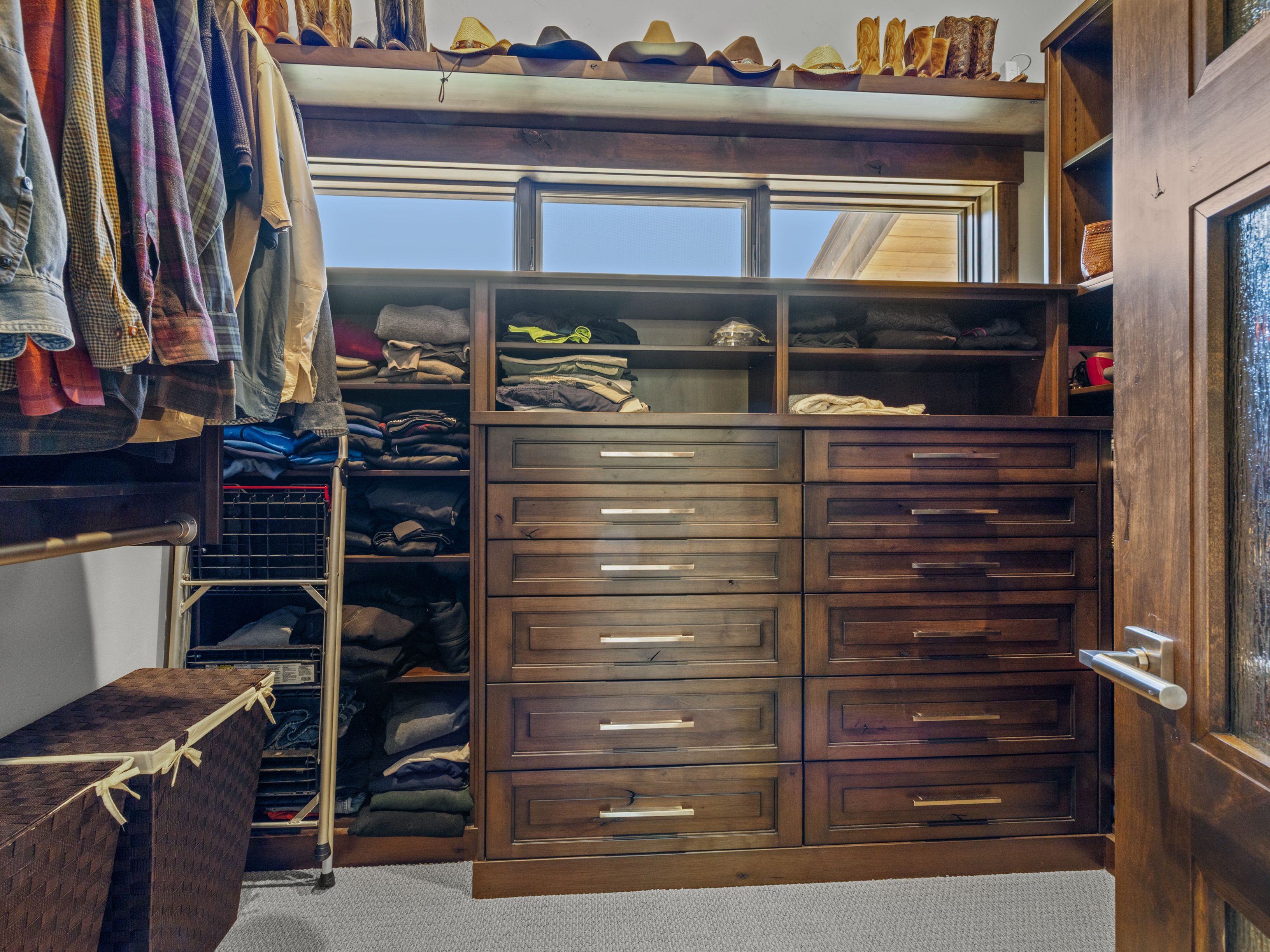 This organized walk-in closet features rich, dark-toned wood cabinetry with extensive drawer storage and open shelving for folded garments. A high shelf above the window displays a collection of cowboy hats and boots, adding a rustic, personalized touch to the space. The room is illuminated by natural light streaming through a horizontal window, highlighting the clean, functional design of the storage system.