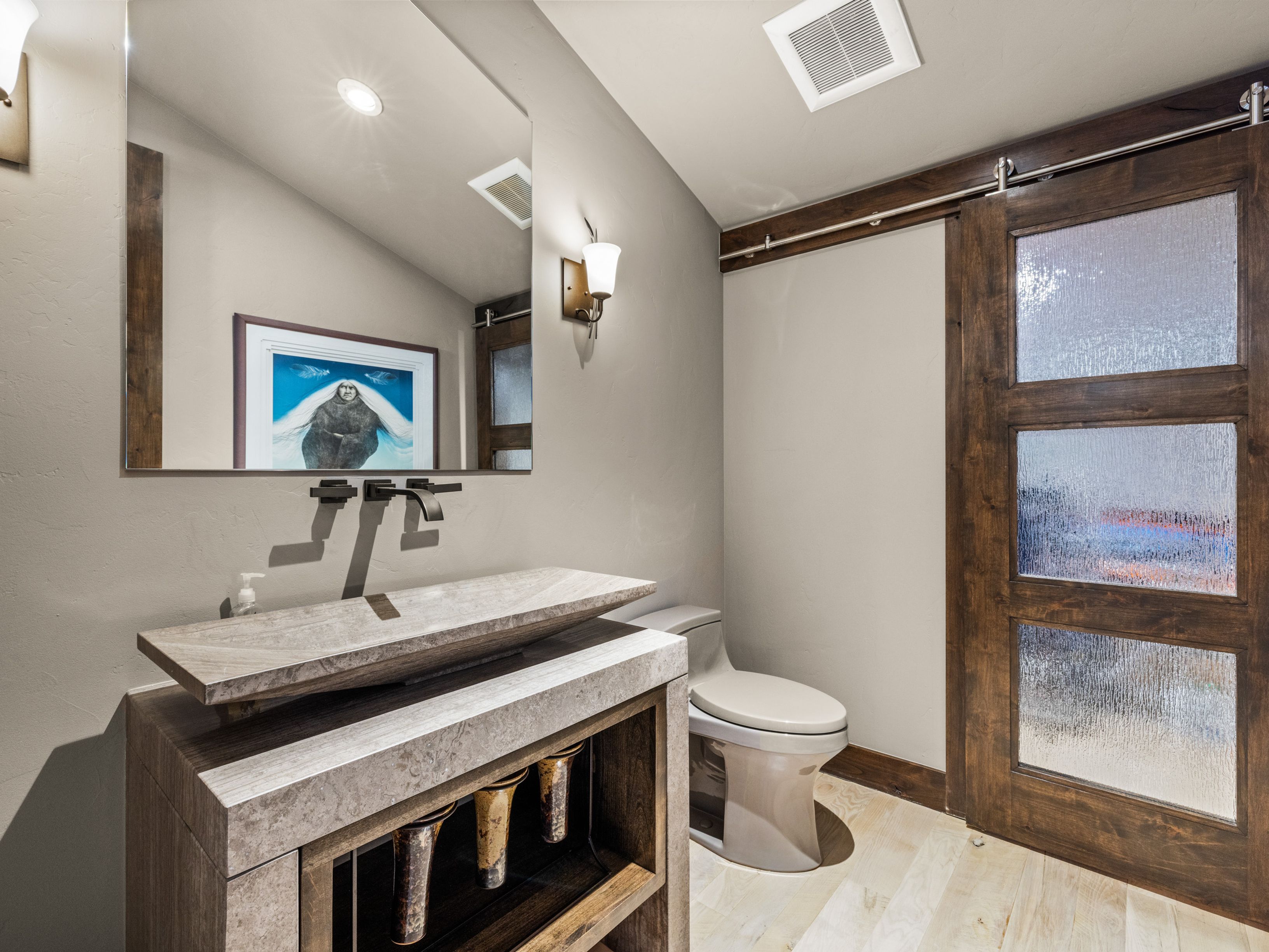 This modern powder room features a unique stone vessel sink set atop a rustic wood and stone vanity. A large, frameless mirror reflects the space, including a wall-mounted light fixture and a piece of framed artwork. To the right, a sliding barn door with textured glass panels adds a rustic touch, while a neutral-toned toilet sits against the wall.