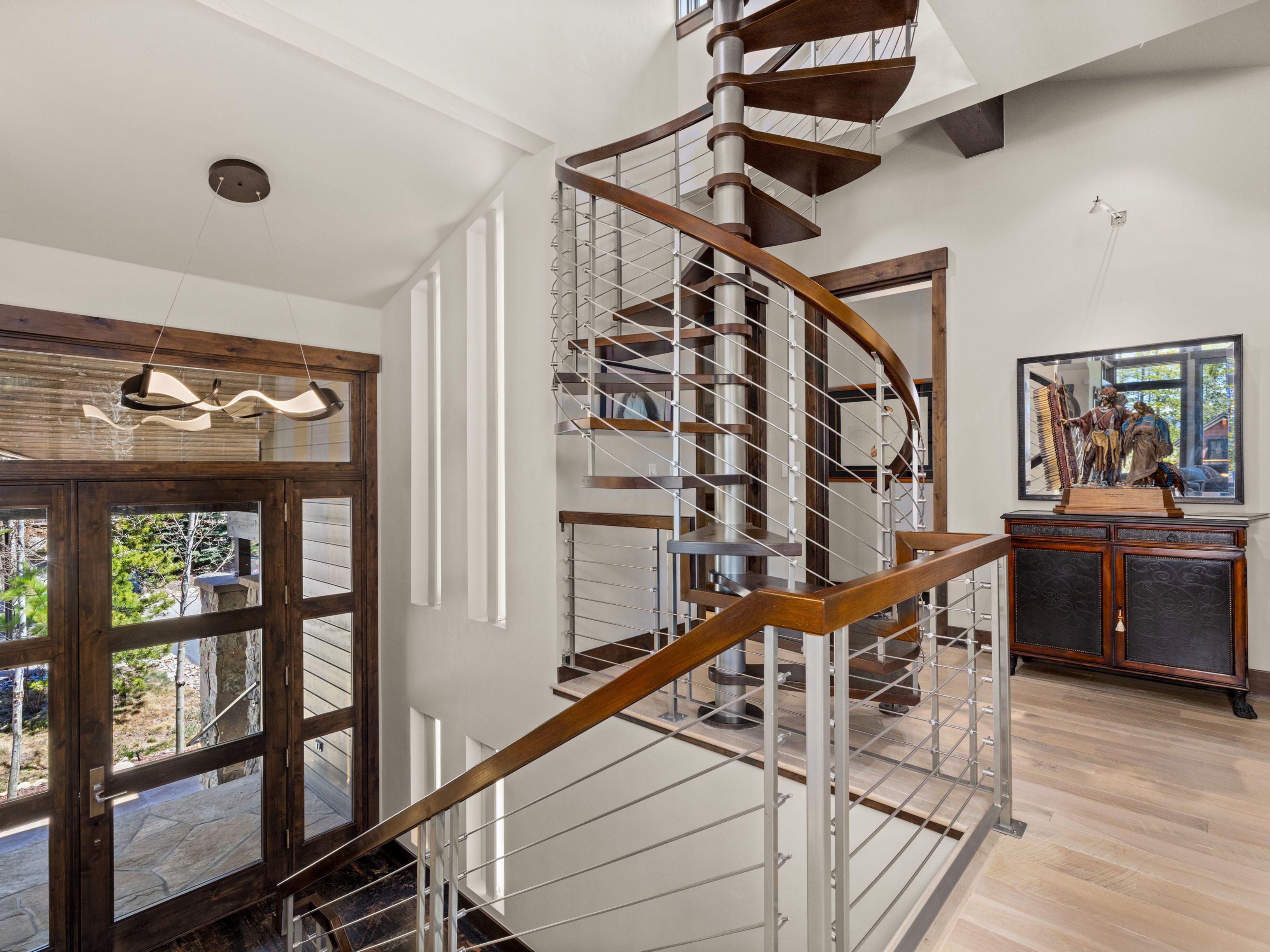 This entryway features a striking spiral staircase with dark wood treads and a sleek metal railing, serving as the focal point of the space. To the left, a large wooden door with glass panels allows natural light to filter in, complemented by a modern, sculptural light fixture overhead. A dark wood cabinet topped with a decorative sculpture sits against the wall, adding a touch of elegance to the light-toned wood flooring and neutral walls.