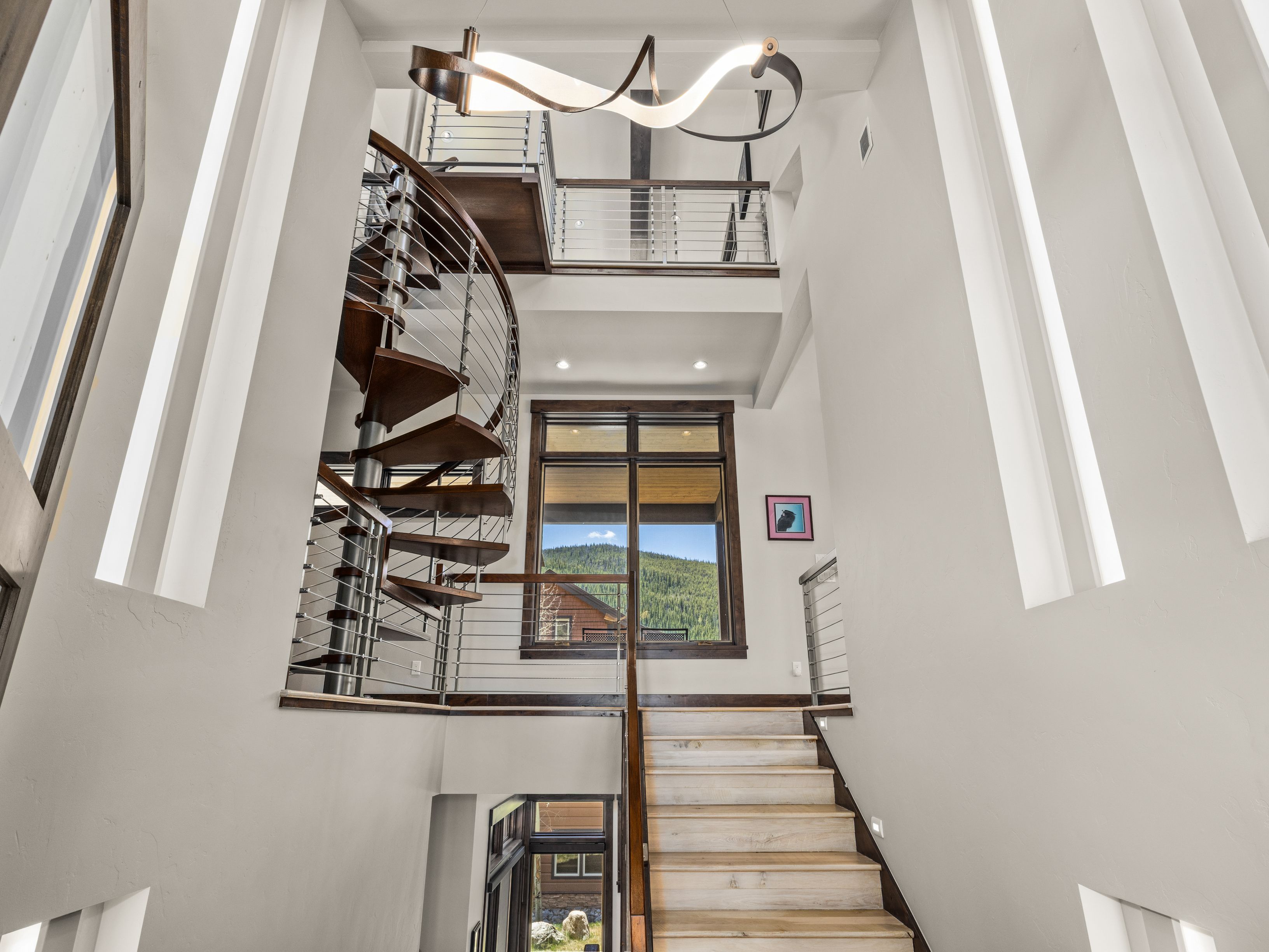 This grand entryway features a striking combination of a spiral staircase with metal railings and a traditional straight staircase with light wood treads. A modern, sculptural light fixture hangs from the high ceiling, drawing the eye upward toward a mezzanine landing. The space is bright and airy, with tall windows that frame a view of the surrounding mountain landscape.