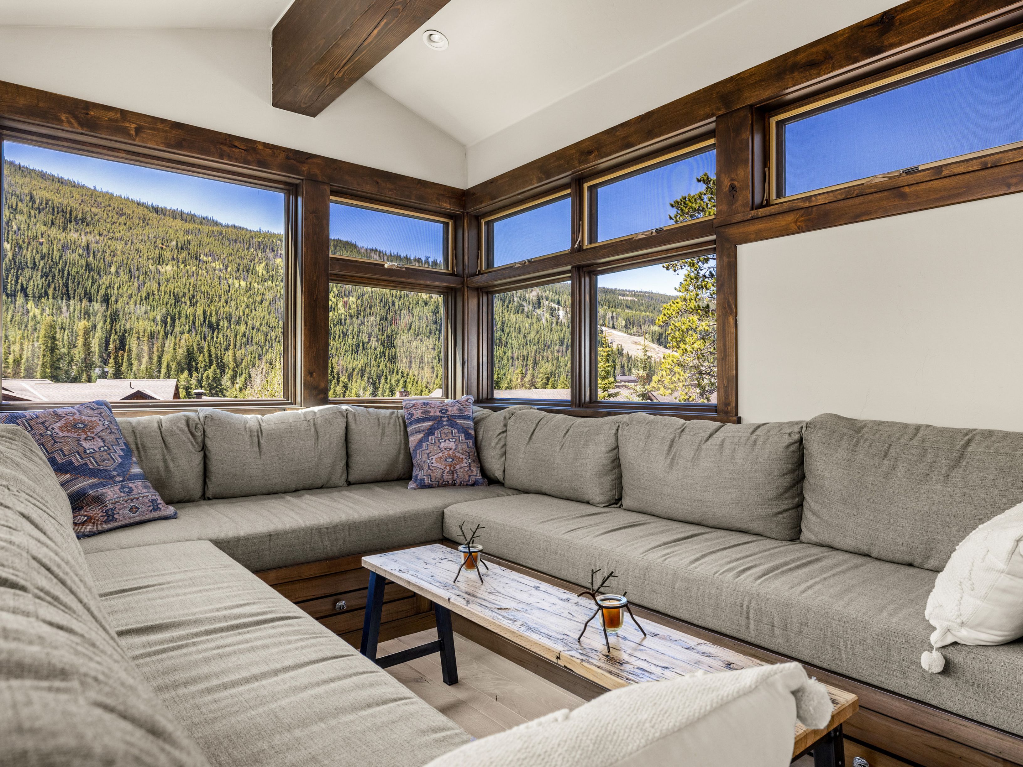 This cozy living area features a large, L-shaped sectional sofa with neutral upholstery and decorative patterned pillows, centered around a rustic wooden coffee table. The space is defined by expansive windows that frame panoramic mountain views, complemented by dark wood trim and a vaulted ceiling with a prominent wooden beam. The overall aesthetic is warm and inviting, perfectly suited for a mountain retreat.