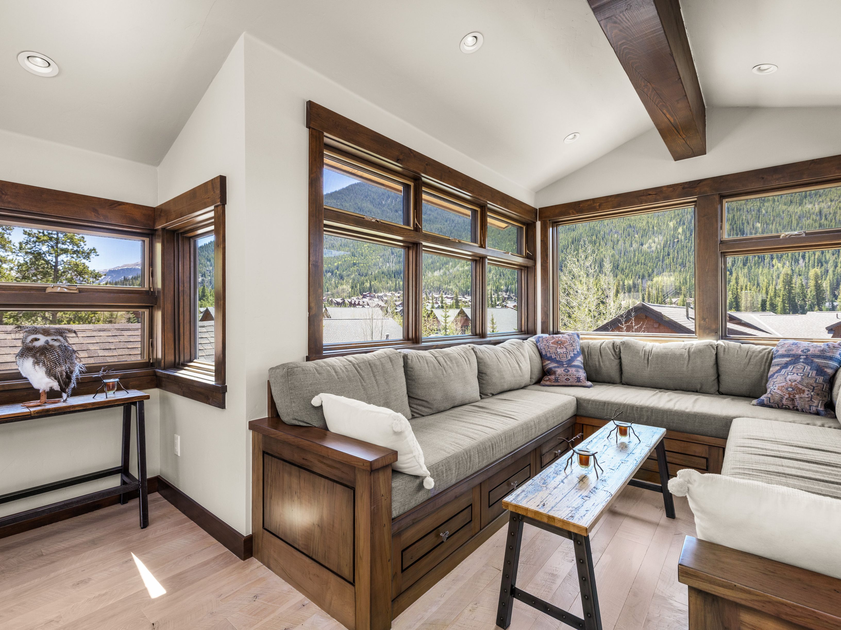This cozy living area features a large, custom-built wooden sectional sofa with plush gray cushions and decorative pillows, positioned to take advantage of the expansive mountain views through large windows. The room is accented by a rustic wooden coffee table, a decorative owl figurine on a side console, and exposed wooden ceiling beams that enhance the mountain-lodge aesthetic. The light wood flooring and neutral walls create a bright, inviting atmosphere perfect for relaxing while looking out at the surrounding forest and residential landscape.