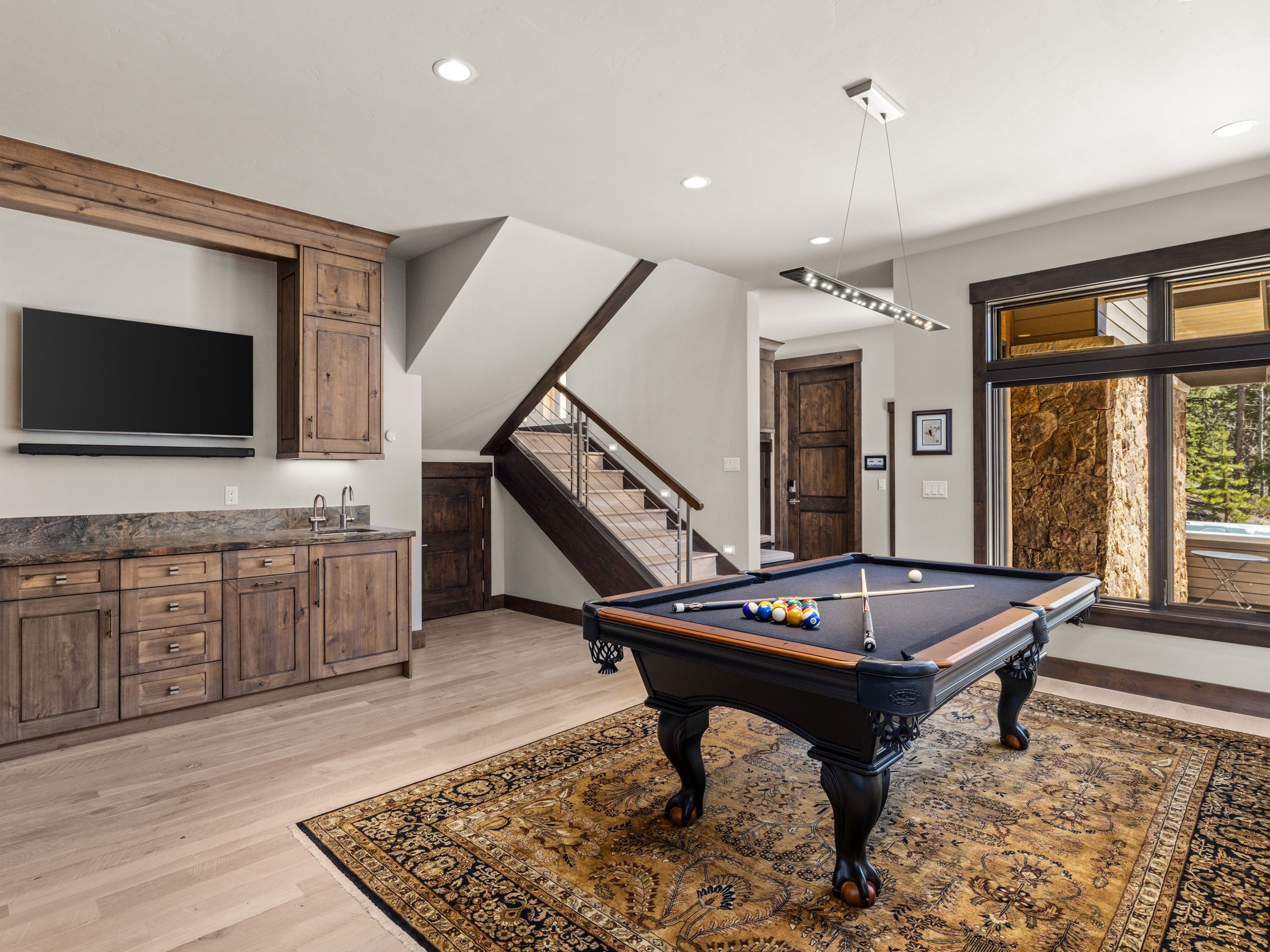 This spacious basement recreation area features a pool table as the central focal point, set atop a large, intricately patterned rug. To the left, a built-in wet bar with dark wood cabinetry and a stone countertop provides a functional entertaining space, while a staircase with a metal railing leads to the upper level. The room is illuminated by recessed lighting and a modern linear pendant fixture, creating a warm and inviting atmosphere for leisure.